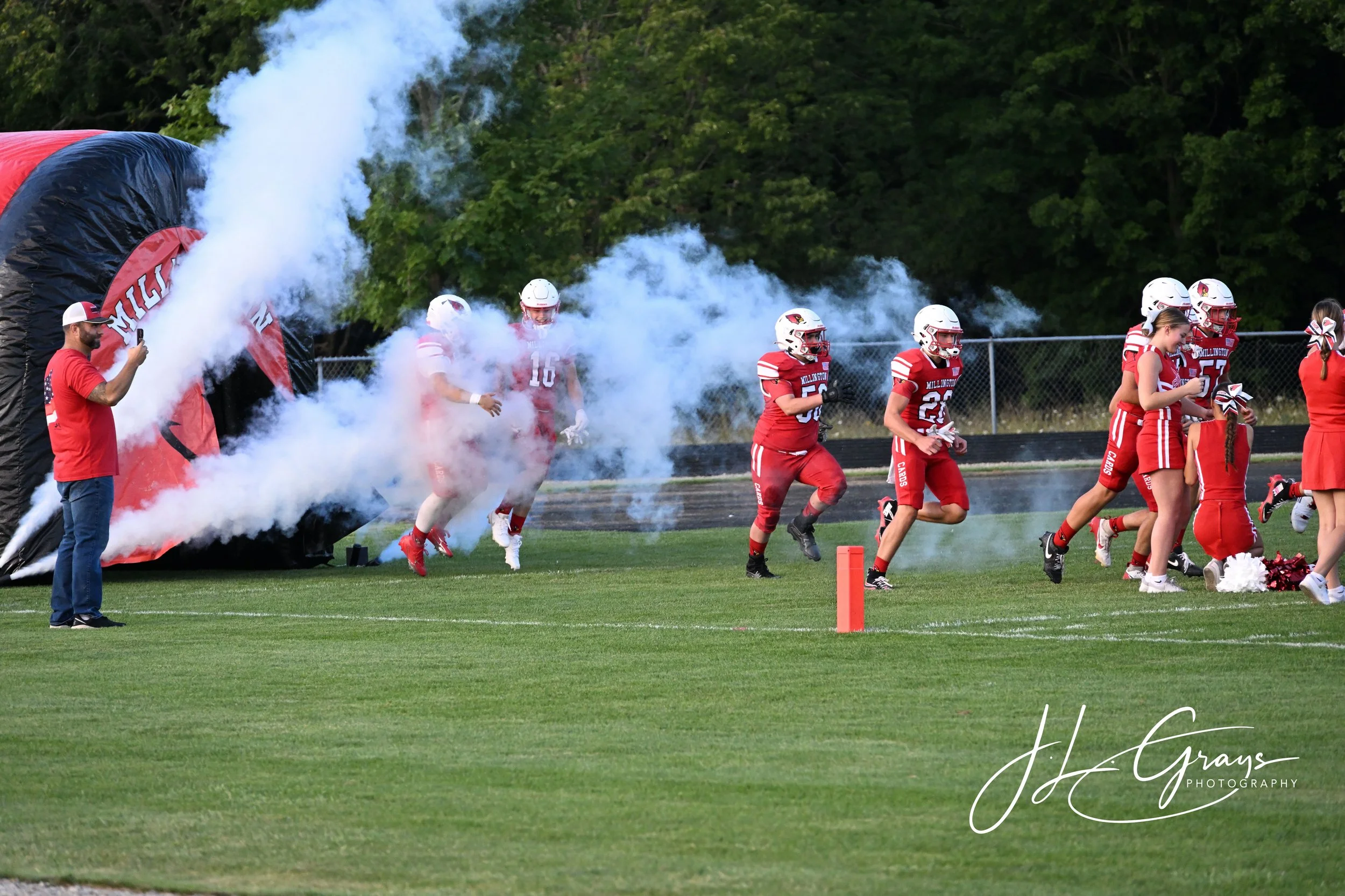 Football players in red uniforms running onto the field through smoke, with a person in a red shirt taking a photo, amid green trees and a chain-link fence.