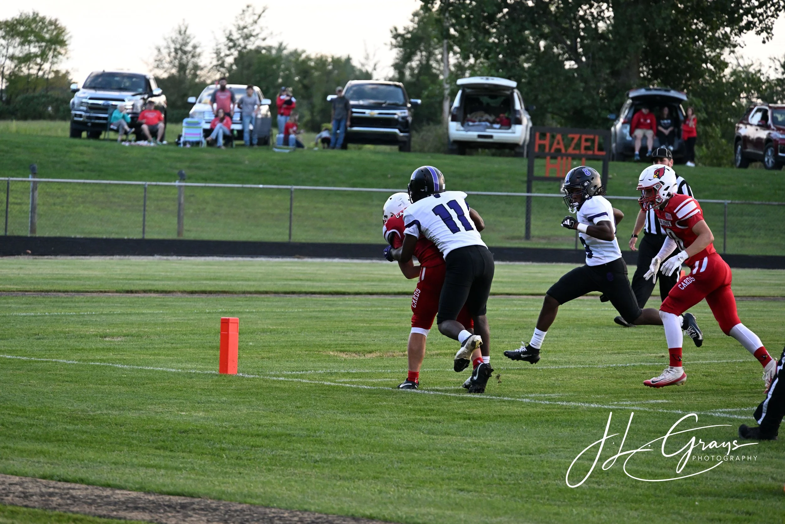 A football game with players in red and white uniforms on the field. A player wearing number 11 in white is holding the ball while being tackled by a player in red. There are spectators sitting on the grass and standing behind a fence, watching the g