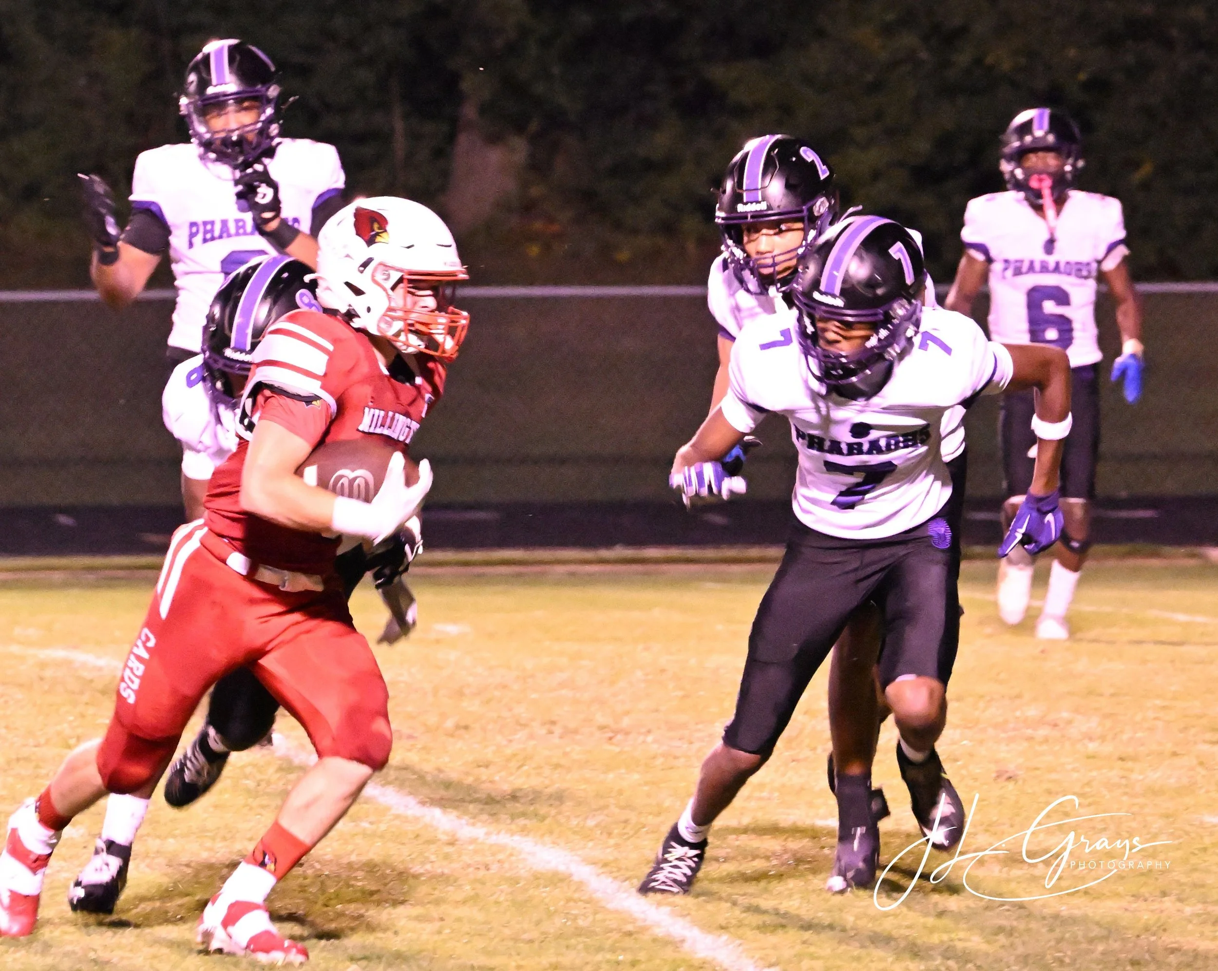A football game at night with players from two teams. One team is wearing white and black uniforms, and the other team is in red uniforms. A player in a red uniform holds a football while running, facing defensive players in white and black uniforms 