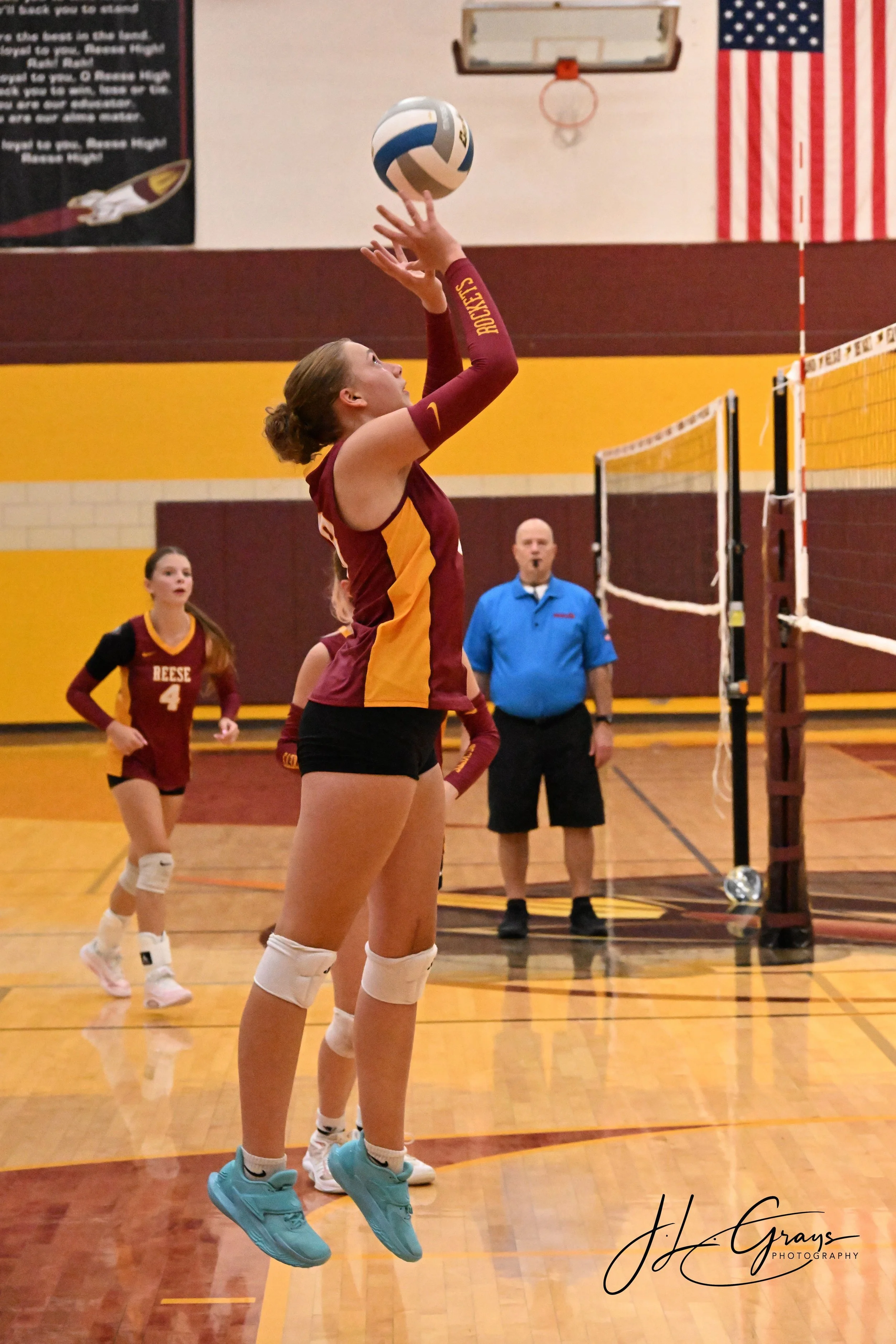 A female volleyball player in a maroon and yellow uniform jumps to set the ball during a volleyball match in a gymnasium with American flag and basketball hoop in the background.