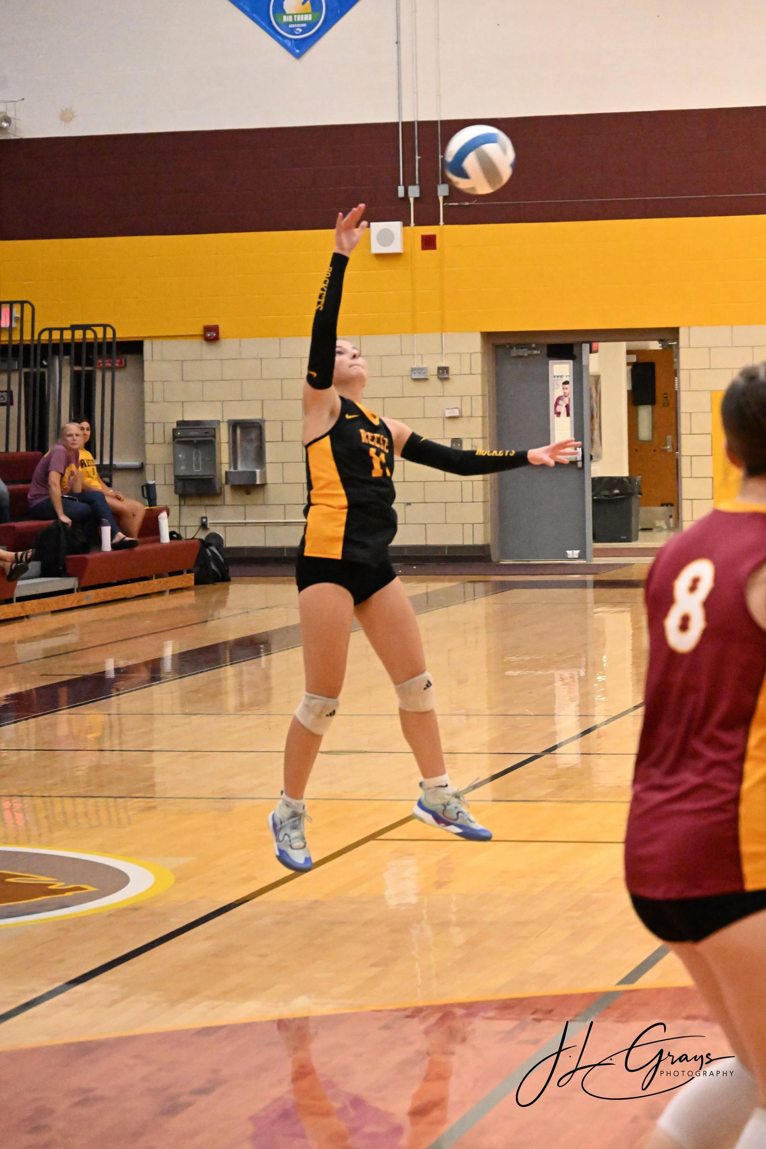A female volleyball player wearing a black and yellow uniform is jumping to hit a volleyball in an indoor gymnasium. She has on knee pads and sports shoes. There are people sitting on benches and a person in a maroon jersey with the number 8 in the f