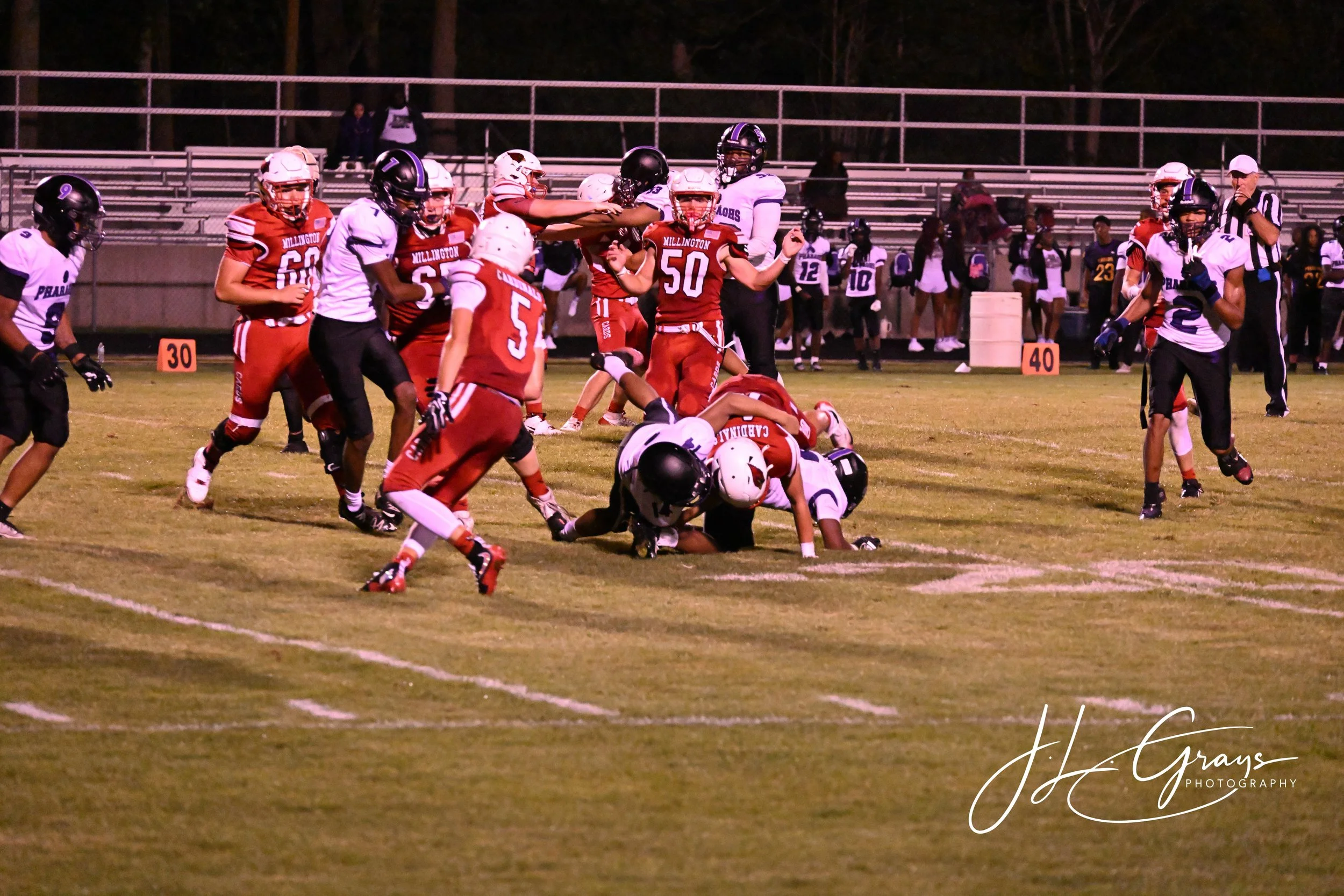A group of high school football players during a game at night, with players from both teams engaging near the ground, some standing and some on the field, with spectators in the background.