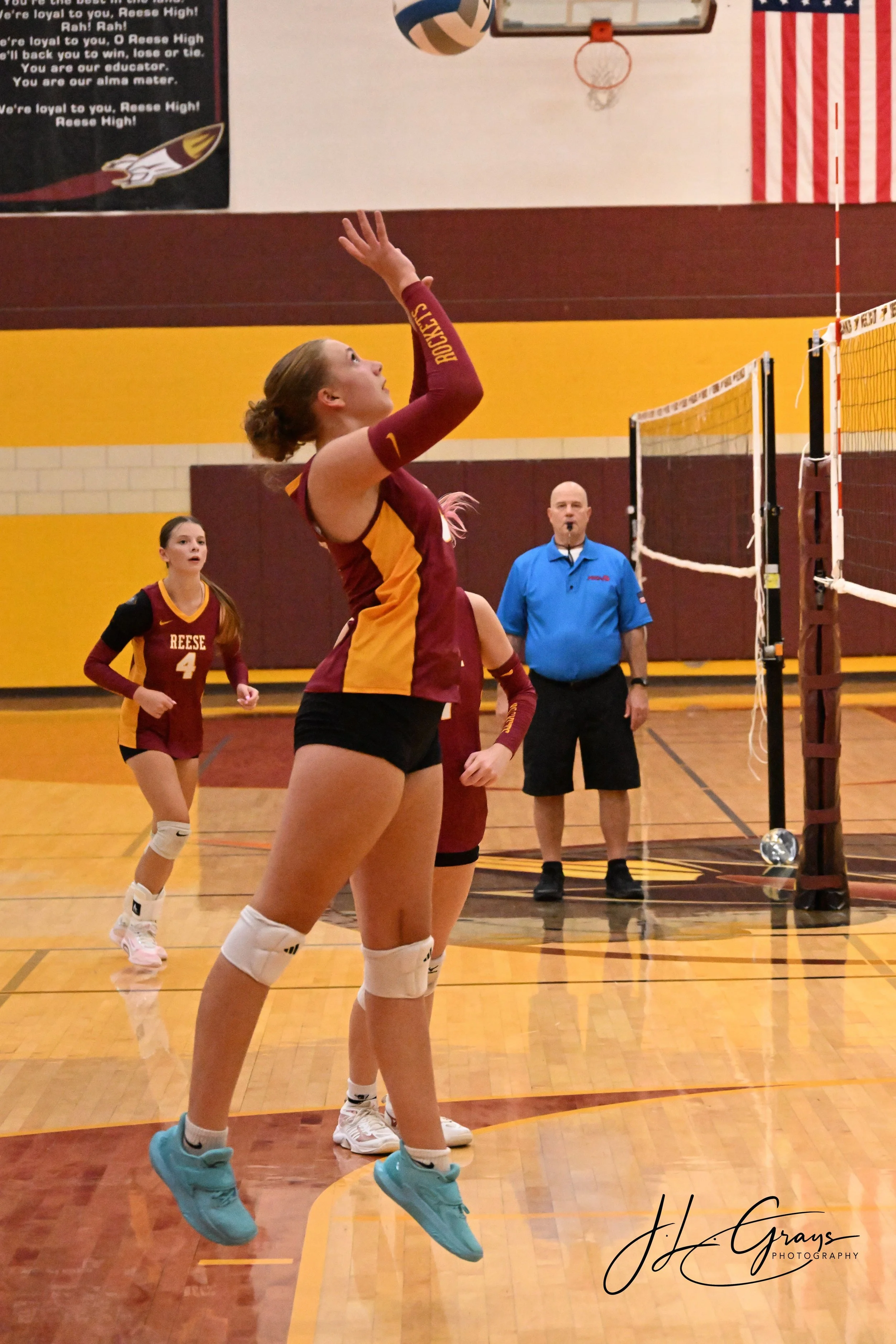 A female volleyball player in a maroon and yellow uniform with "REese" on the front jumps to hit the volleyball. She wears turquoise shoes, knee pads, and has her hair tied back. Two other players in similar uniforms are visible in the background, al
