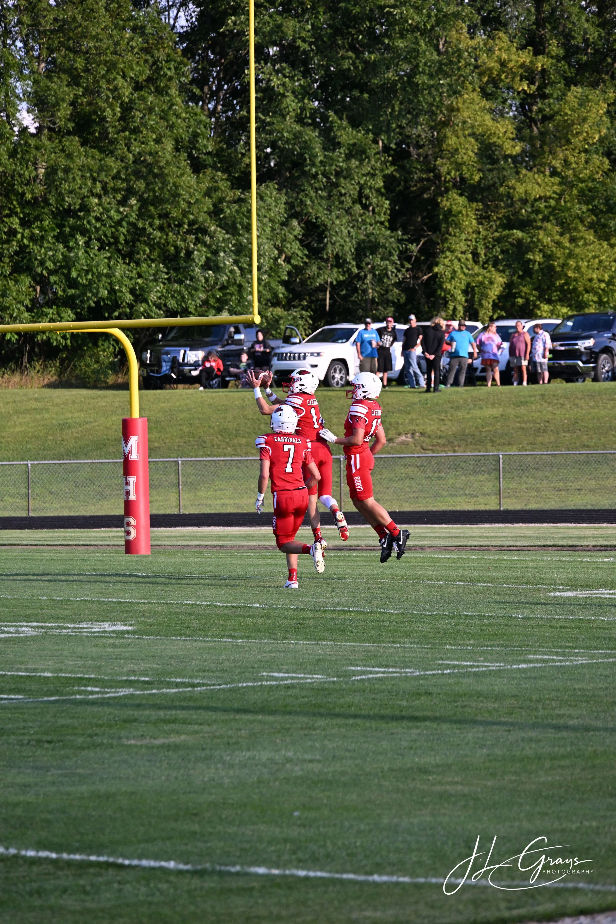 American football players in red uniforms celebrating a touchdown in a football field, with spectators and parked cars in the background.
