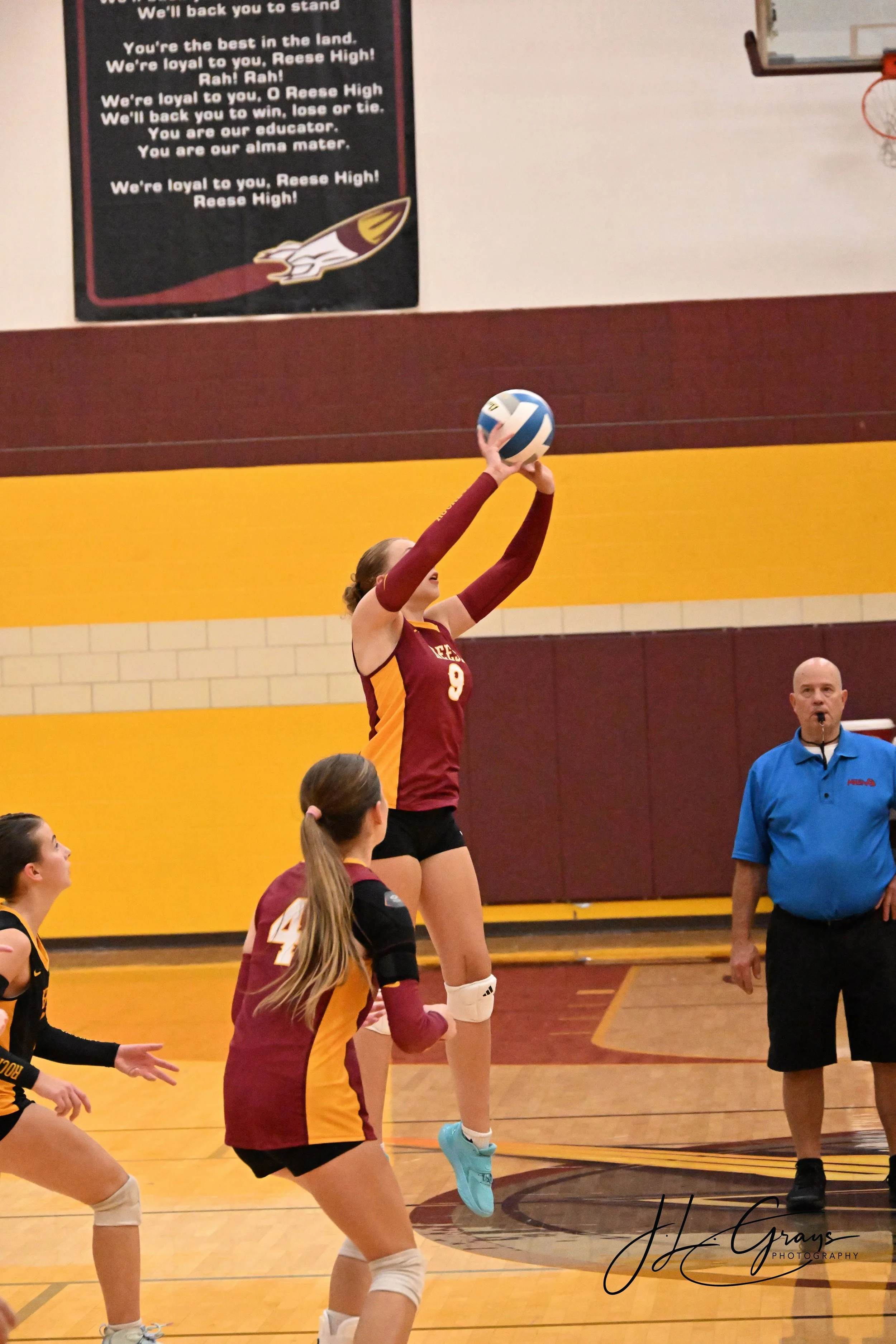 A volleyball player in a maroon and yellow uniform jumps to hit the ball during a game in a gymnasium. Other players are on the court, and a referee stands nearby. A poster with a rallying chant and a logo is on the wall in the background.