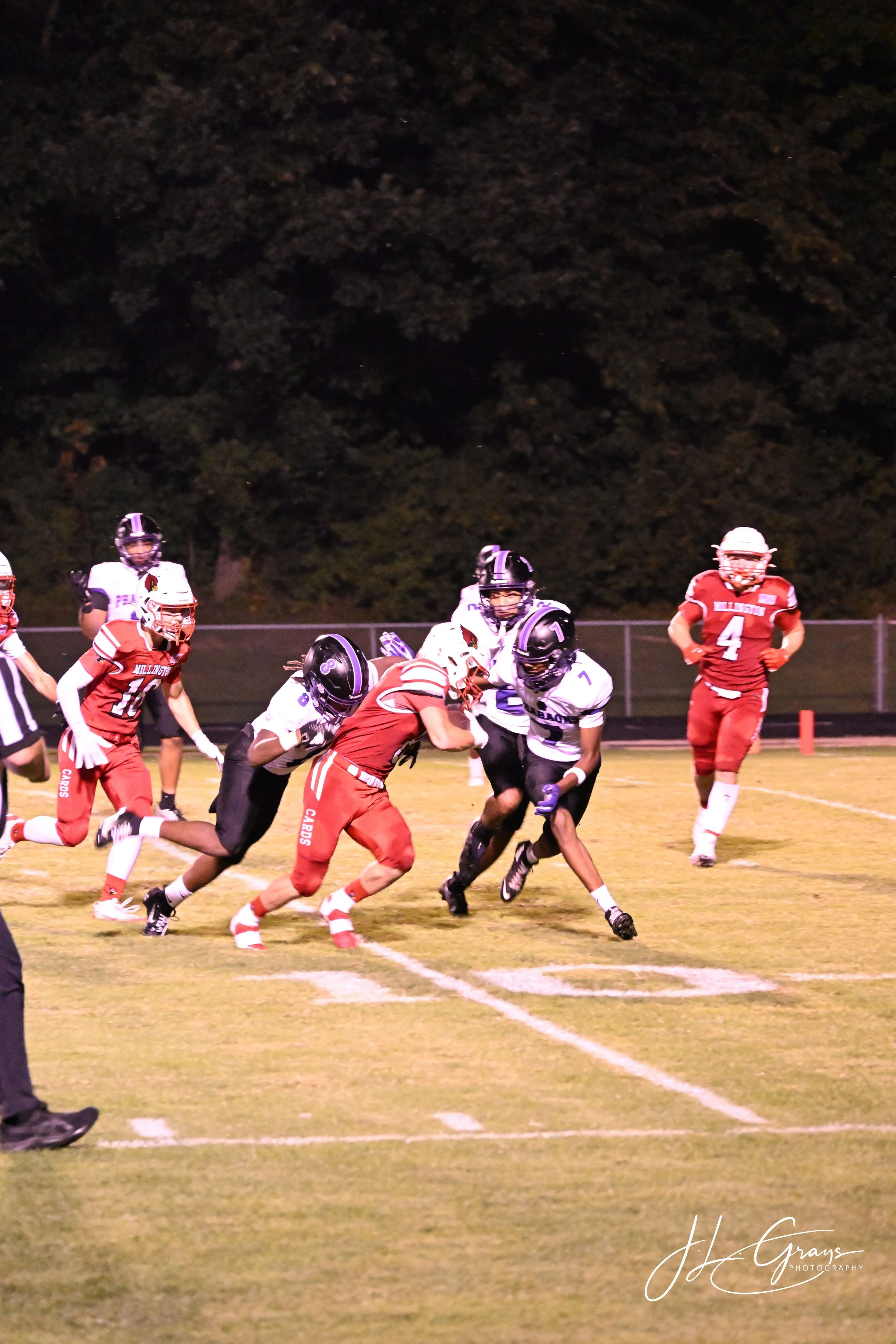 A high school football game at night with players on the field. The team in red uniforms is playing against the team in white and black uniforms. The players are engaged in a play, and there is a referee on the left side. A dark, tree-filled backgrou