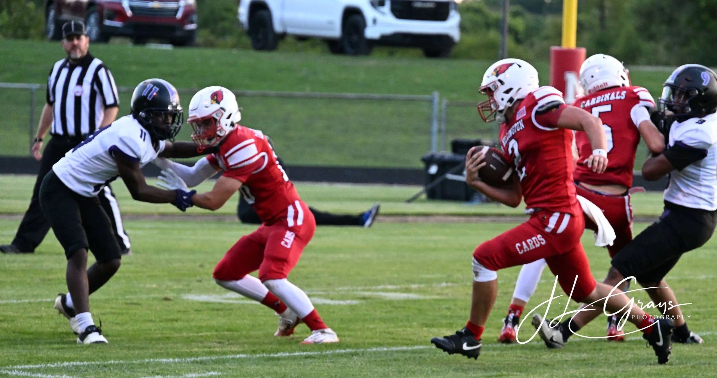 A football game with players in red and black uniforms. One player in red holding the football, running, while a player in a black and white uniform attempts to tackle. An official stands nearby on the field with a green grass playing surface in the 