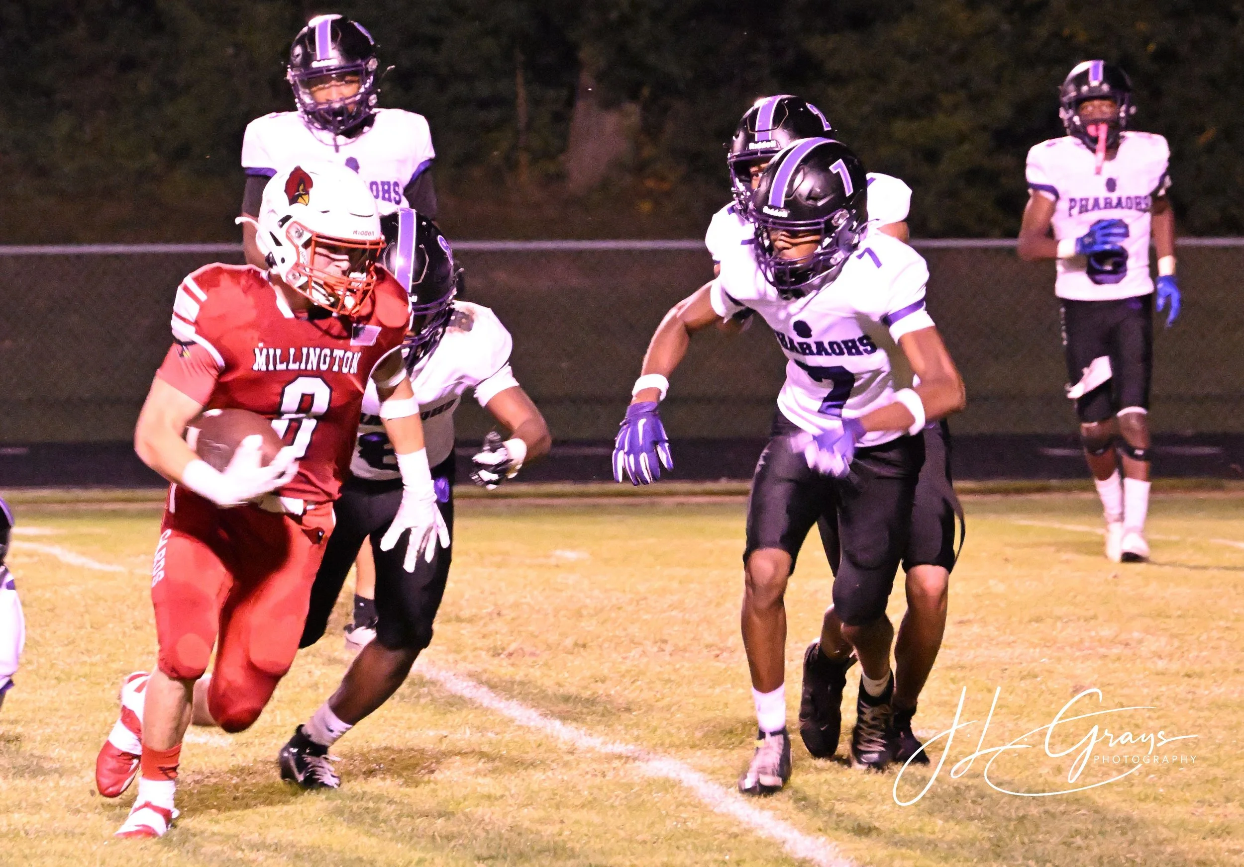 Young football player in red uniform carrying a football, running while others in white uniforms approach him during night game.
