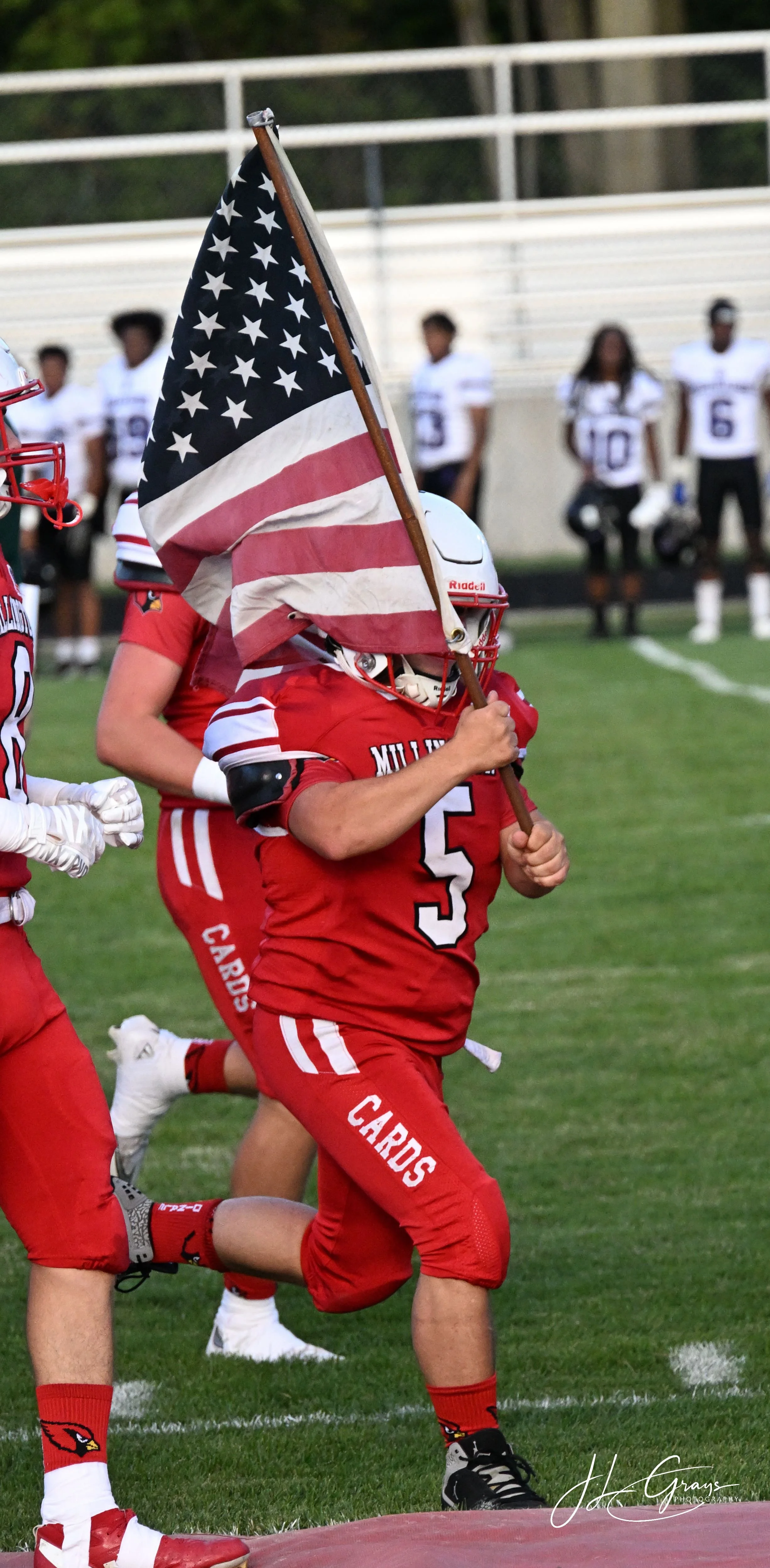 A football player in a red uniform, number 5, holding an American flag, running on a football field during a game. Other players in the background are standing on the sidelines.