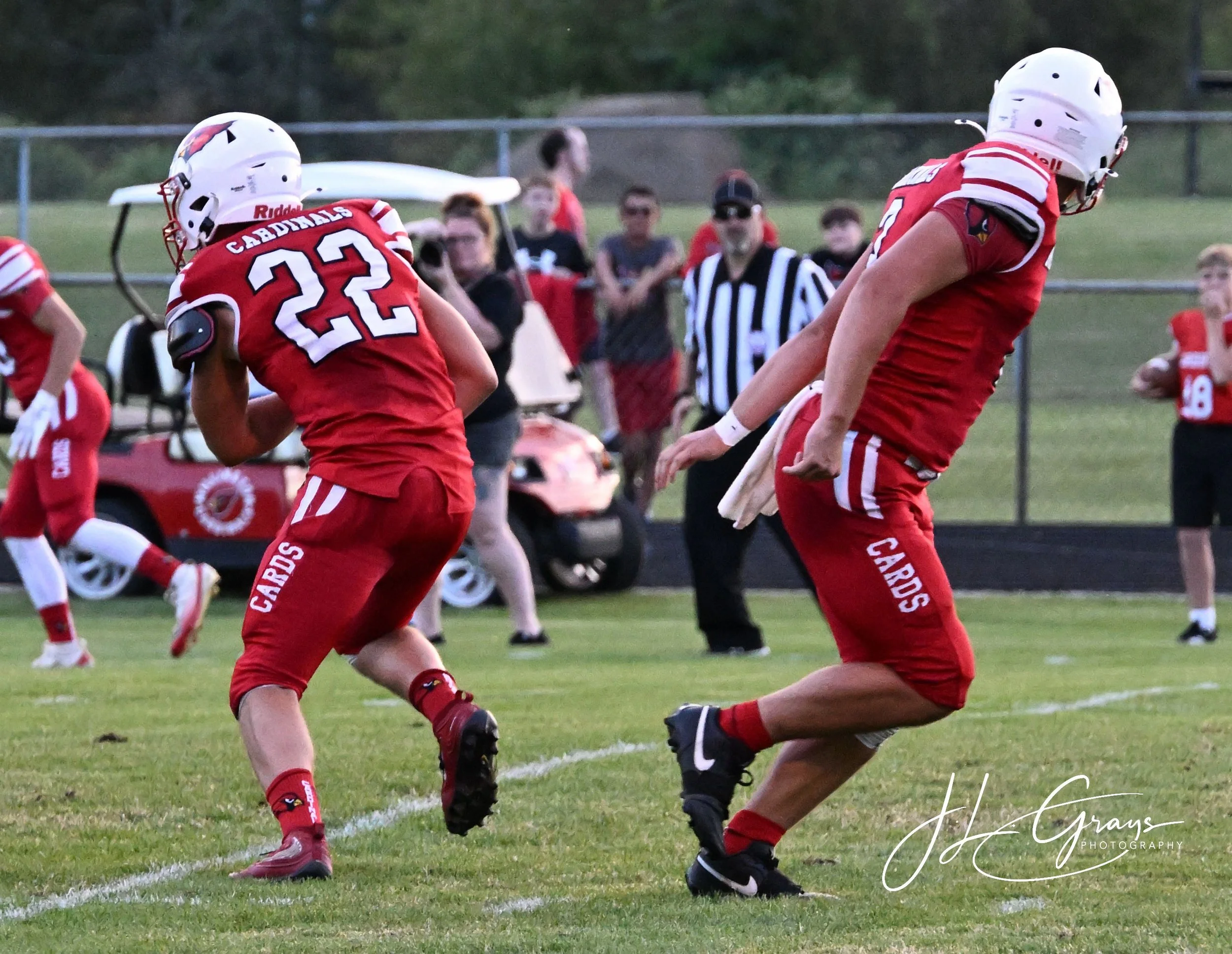 Two young football players in red uniforms and white helmets are playing on a grassy field; one player is running while the other is preparing to block or tackle.