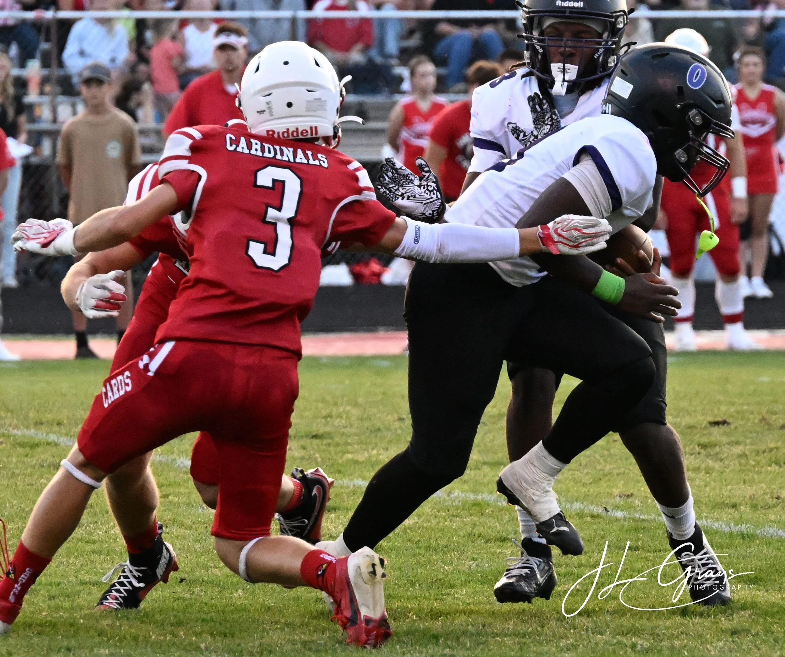 Two football players, one in a red jersey and the other in a white jersey, engaging in a play on the field. The player in white is holding the football while the player in red is reaching to tackle him. Spectators watch from the stands in the backgro