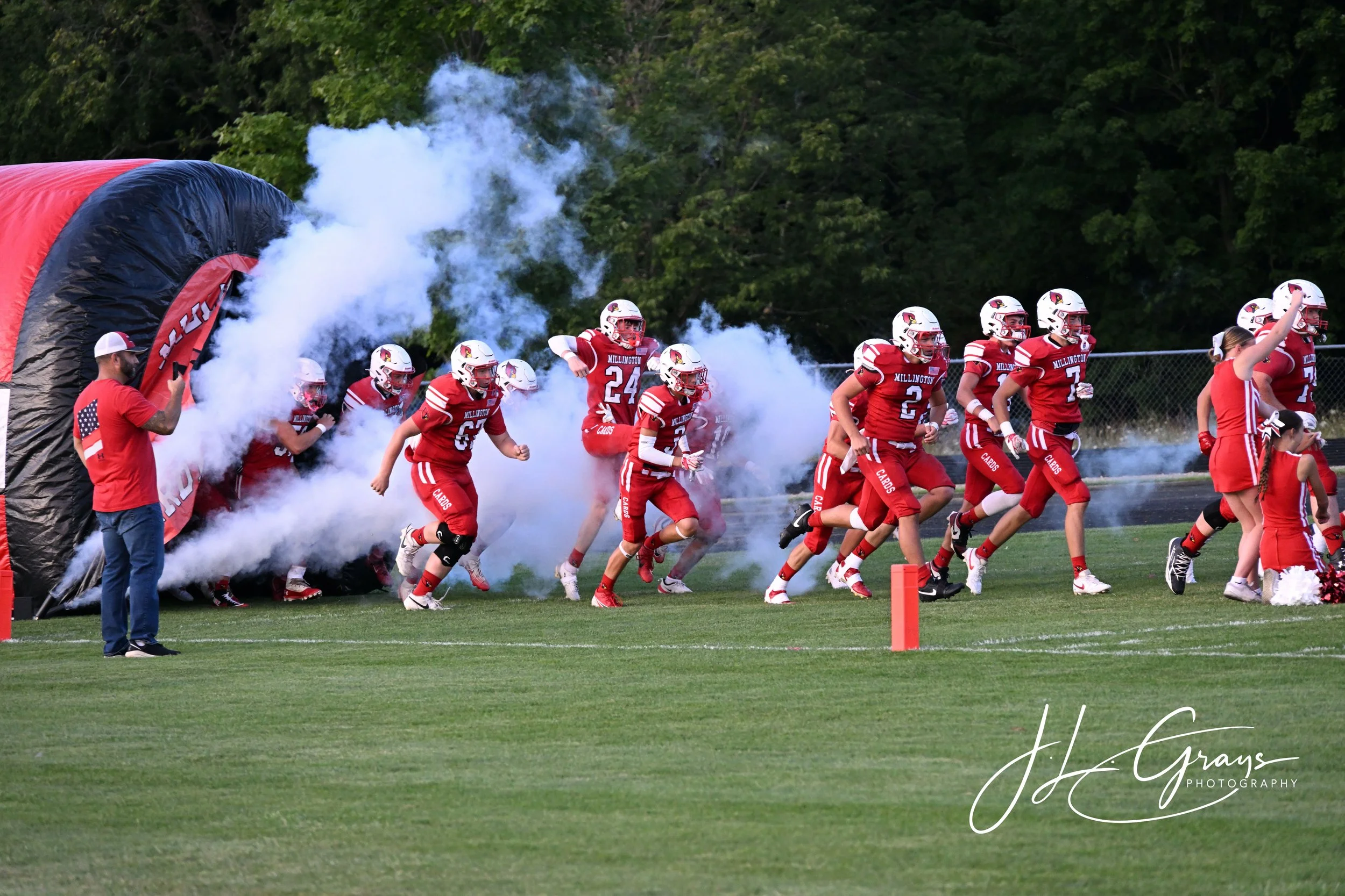 High school football team running onto the field through a smoke tunnel during a game. Players are wearing red uniforms with white helmets and are followed by a person holding a flag. The background features green trees and a chain-link fence.