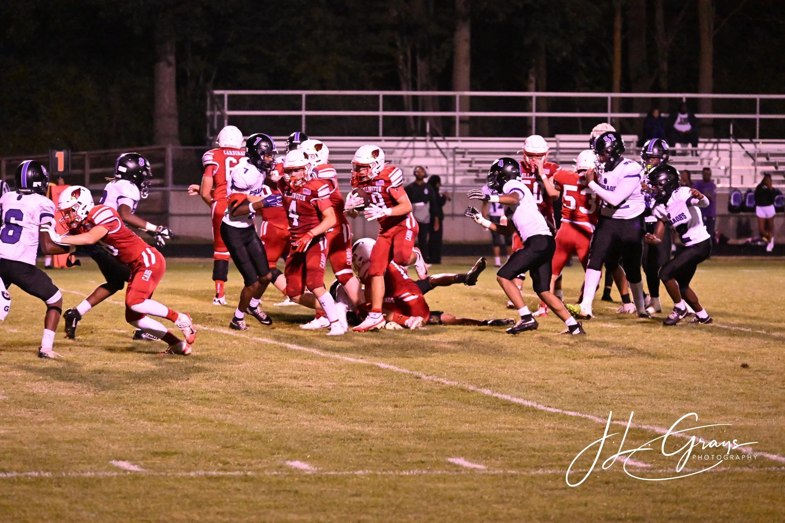 A nighttime high school football game with players in red and white uniforms engaging in a play on the field, with spectators in the background.