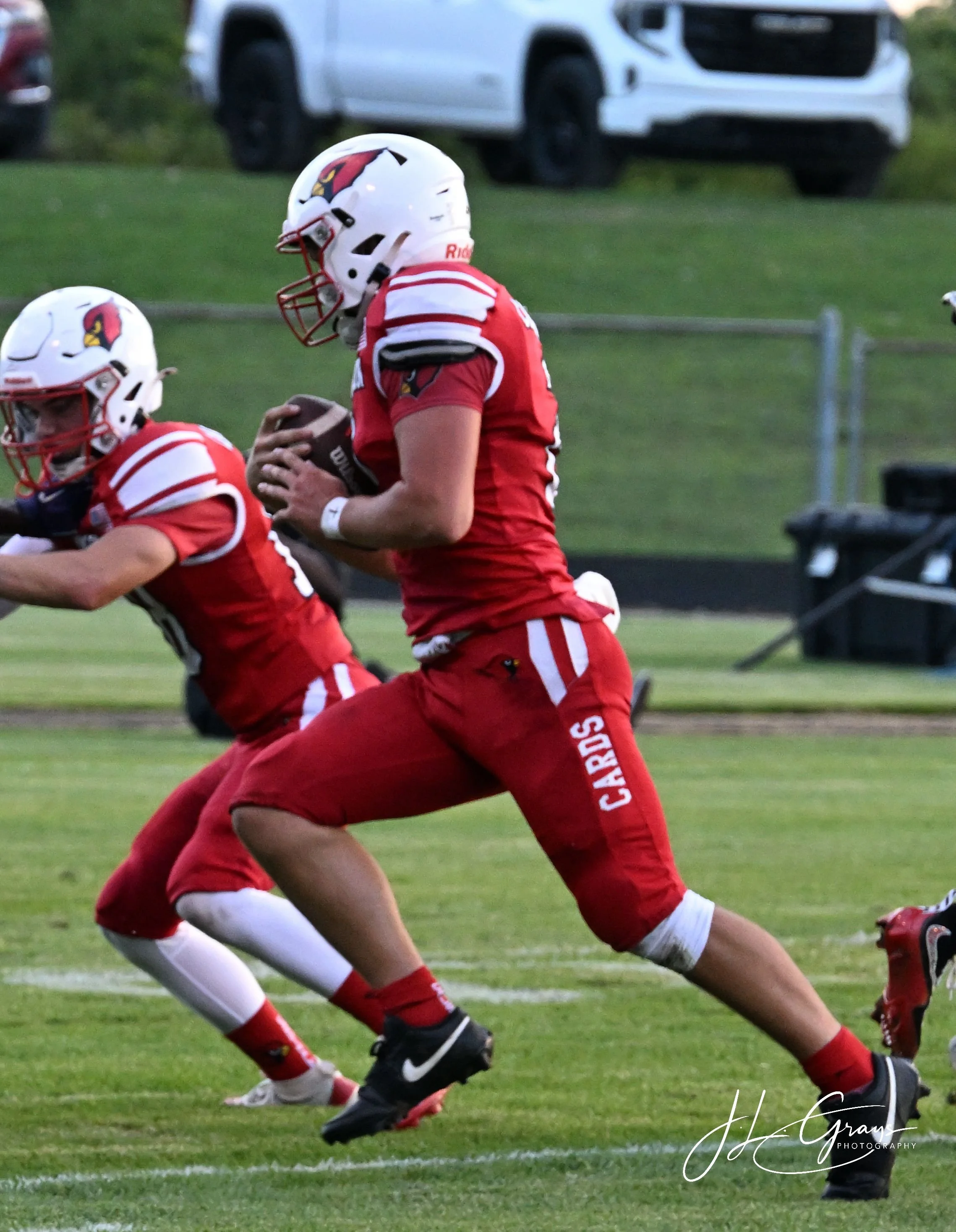 American football players in red uniforms and helmets, one holding a football, engaging in a game on a grassy field.