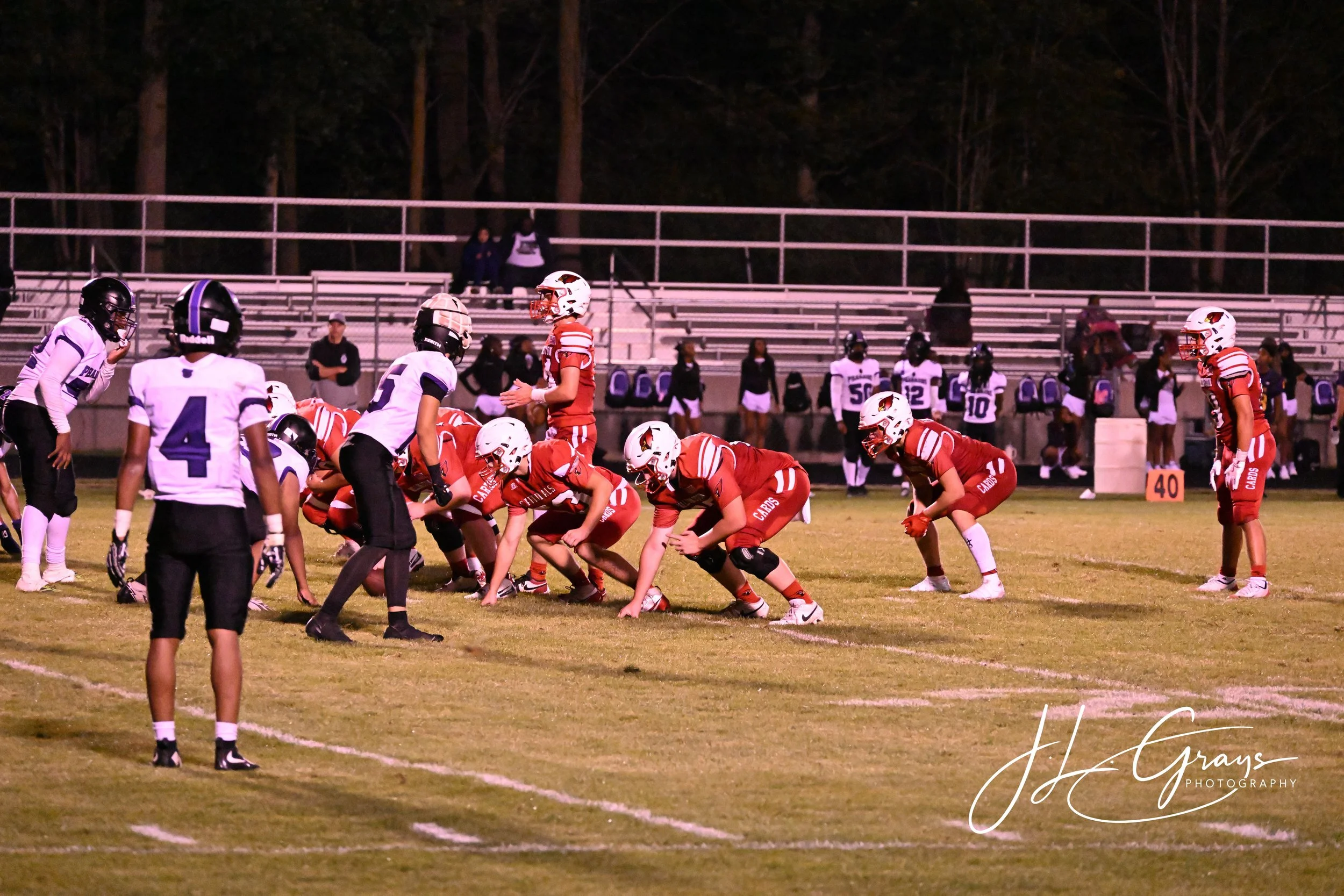 A nighttime football game with players lined up on the field, wearing uniforms and helmets, with spectators in the bleachers and a wooded background.