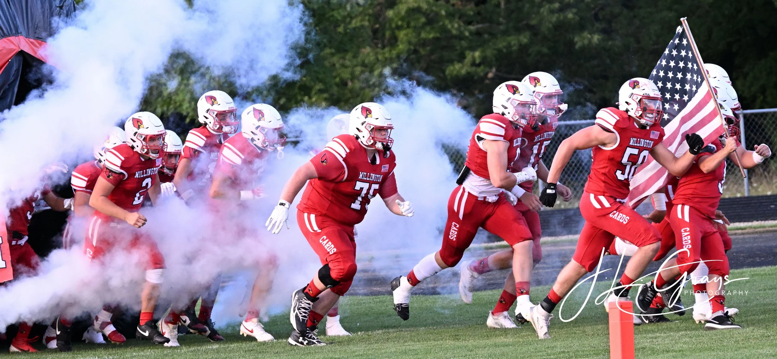High school football team in red uniforms running onto the field through smoke, with one player carrying an American flag.