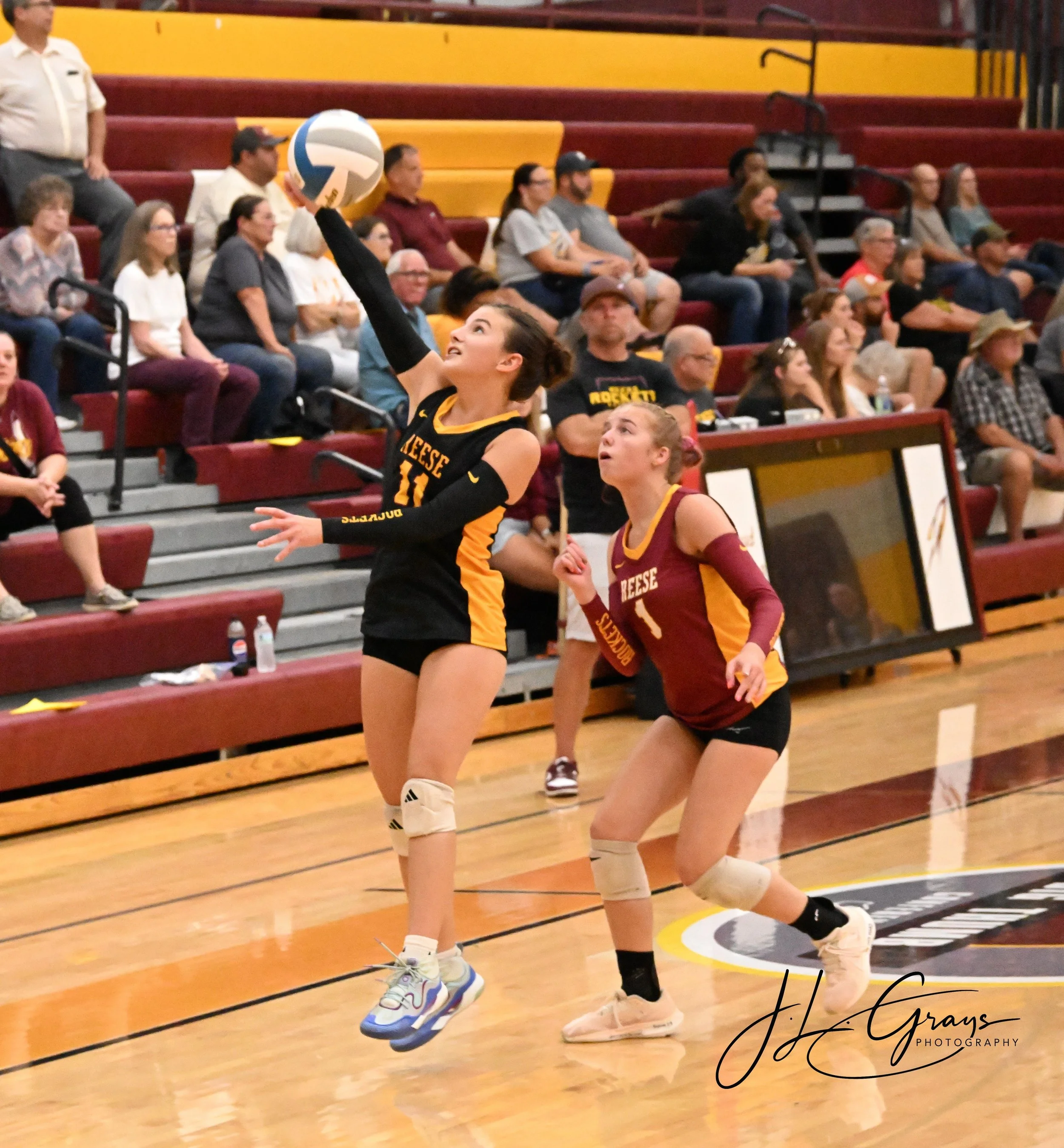 Two female volleyball players in mid-air during a game, with an audience in the background sitting on bleachers, in a gymnasium with wood floors and yellow and maroon walls.