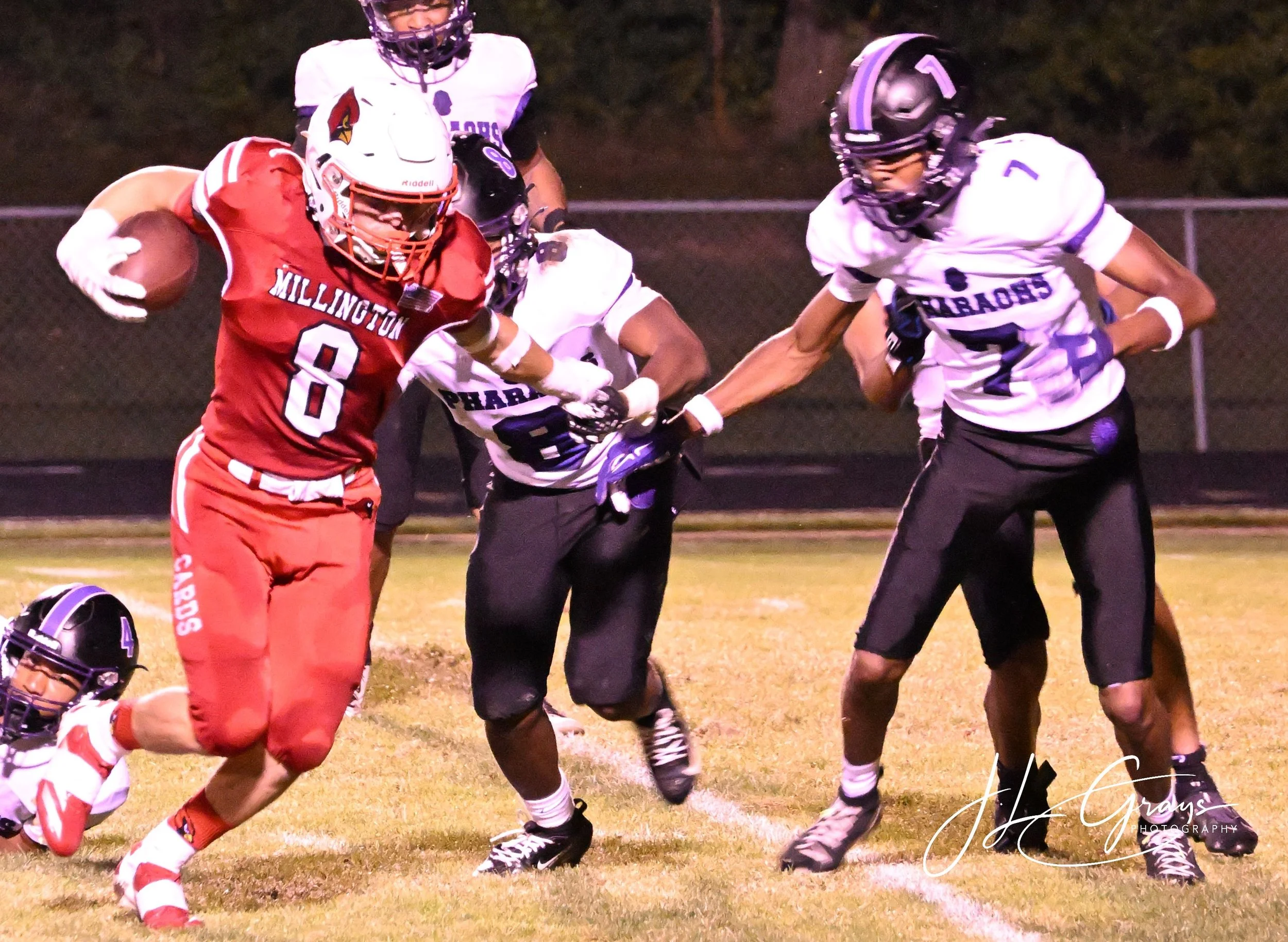 Football players in action on the field during a game at night. One player in red is holding the football, while players in black and white are trying to stop him.
