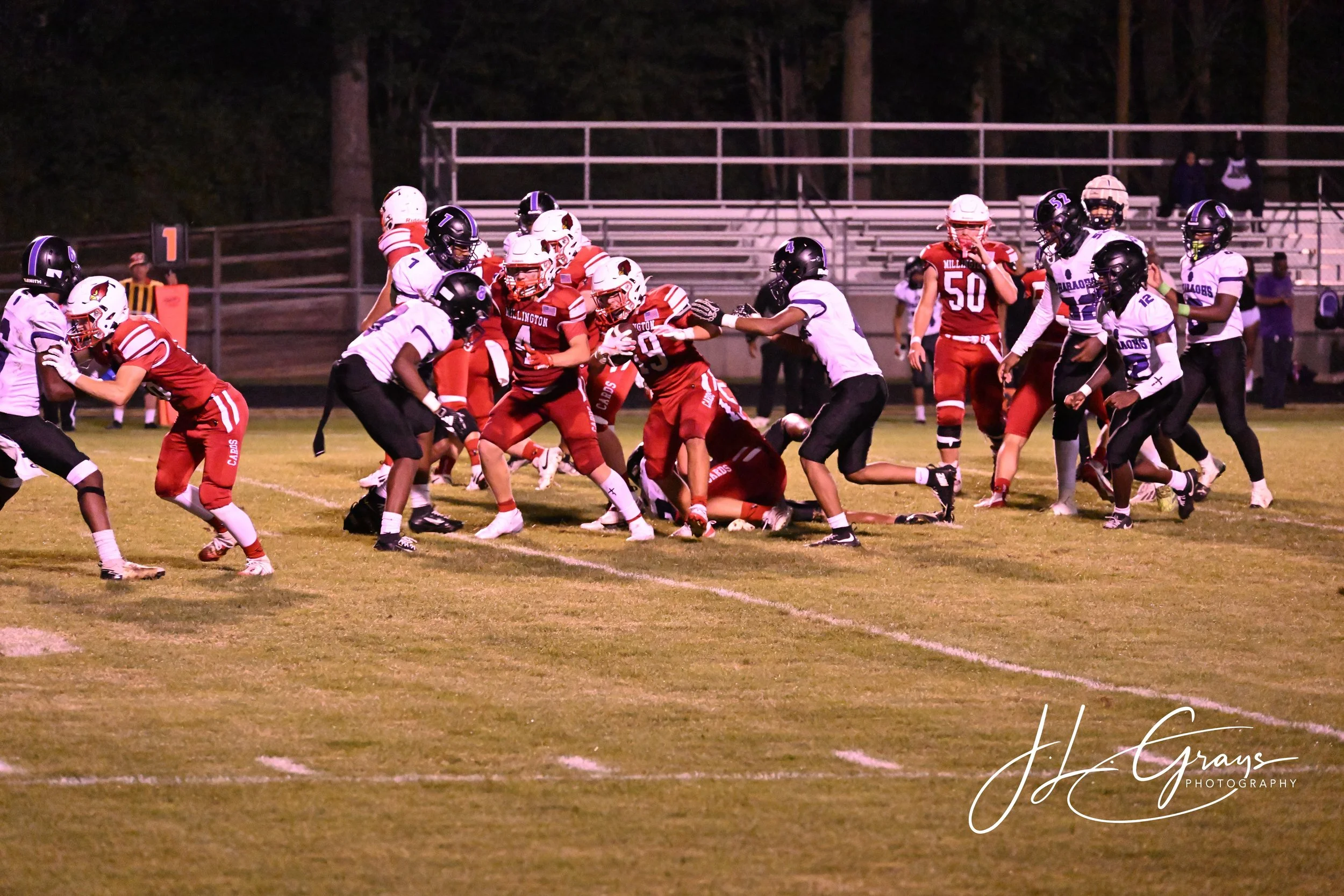 Nighttime football game with players from two teams, wearing red and white or black and white uniforms, engaged in a play on the field surrounded by empty bleachers.
