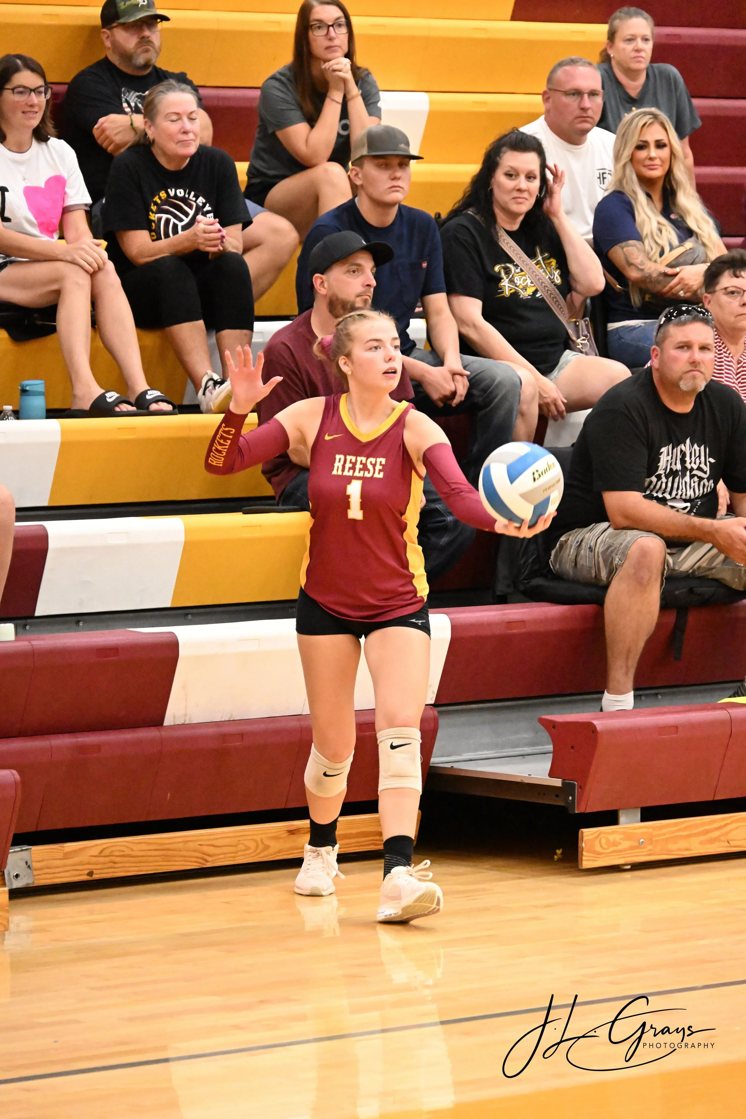 A female volleyball player wearing a maroon and yellow uniform with the name 'Reese' and the number 1, standing on a gymnasium court, holding a volleyball, with spectators seated in the background.