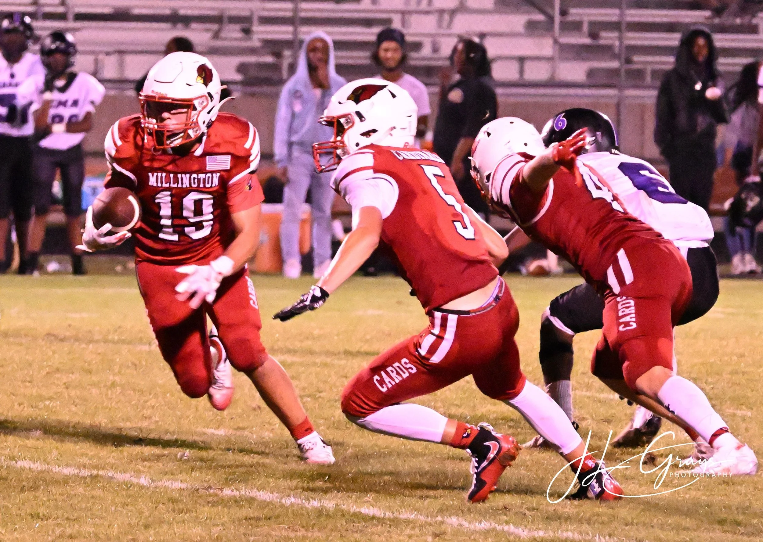 Young football players in red and white uniforms play on a grassy field during a game, with spectators watching from the bleachers in the background.