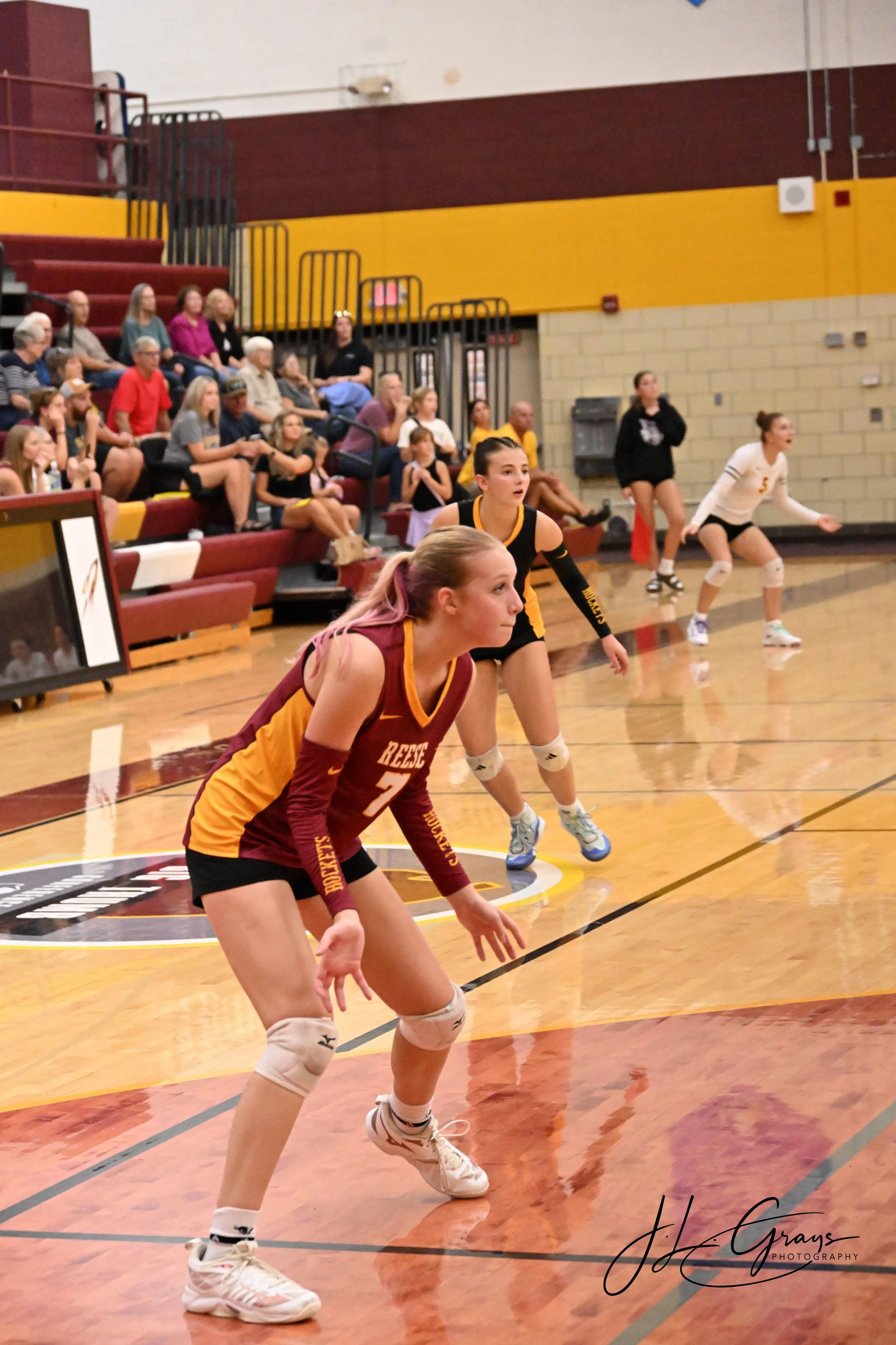 A volleyball game taking place indoors with players on the court and spectators watching from the bleachers.