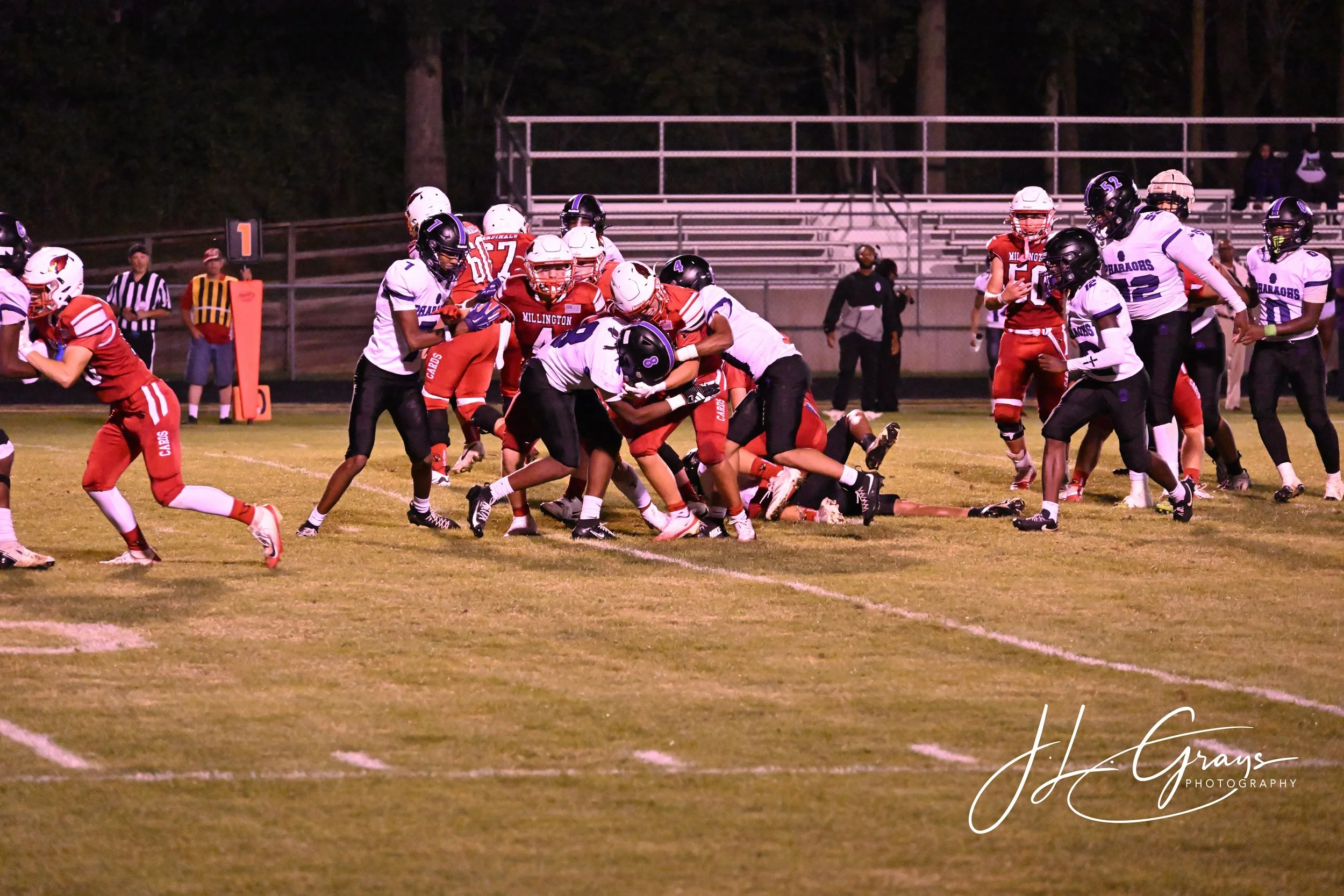 A group of football players engaged in a play on a football field during a game at night. Players are wearing uniforms, helmets, and protective gear, with some in red and others in white and black. The scene depicts an intense moment of tackling or b
