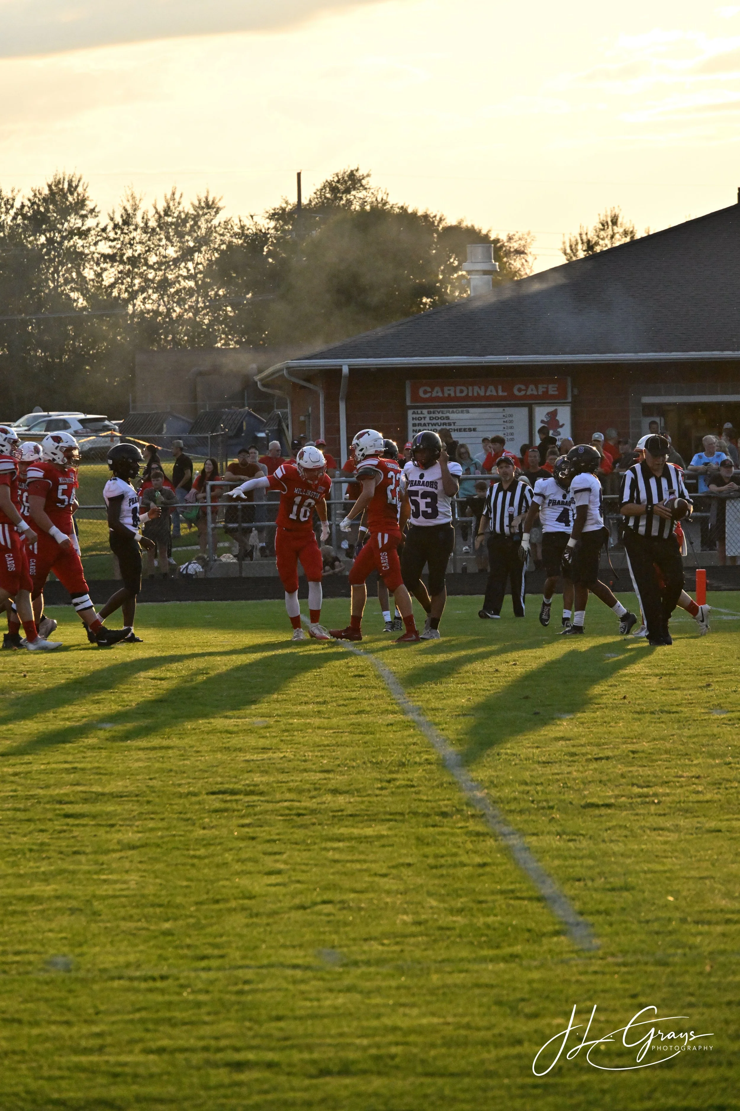 High school football players on the field during a game at sunset, with spectators and a building in the background.