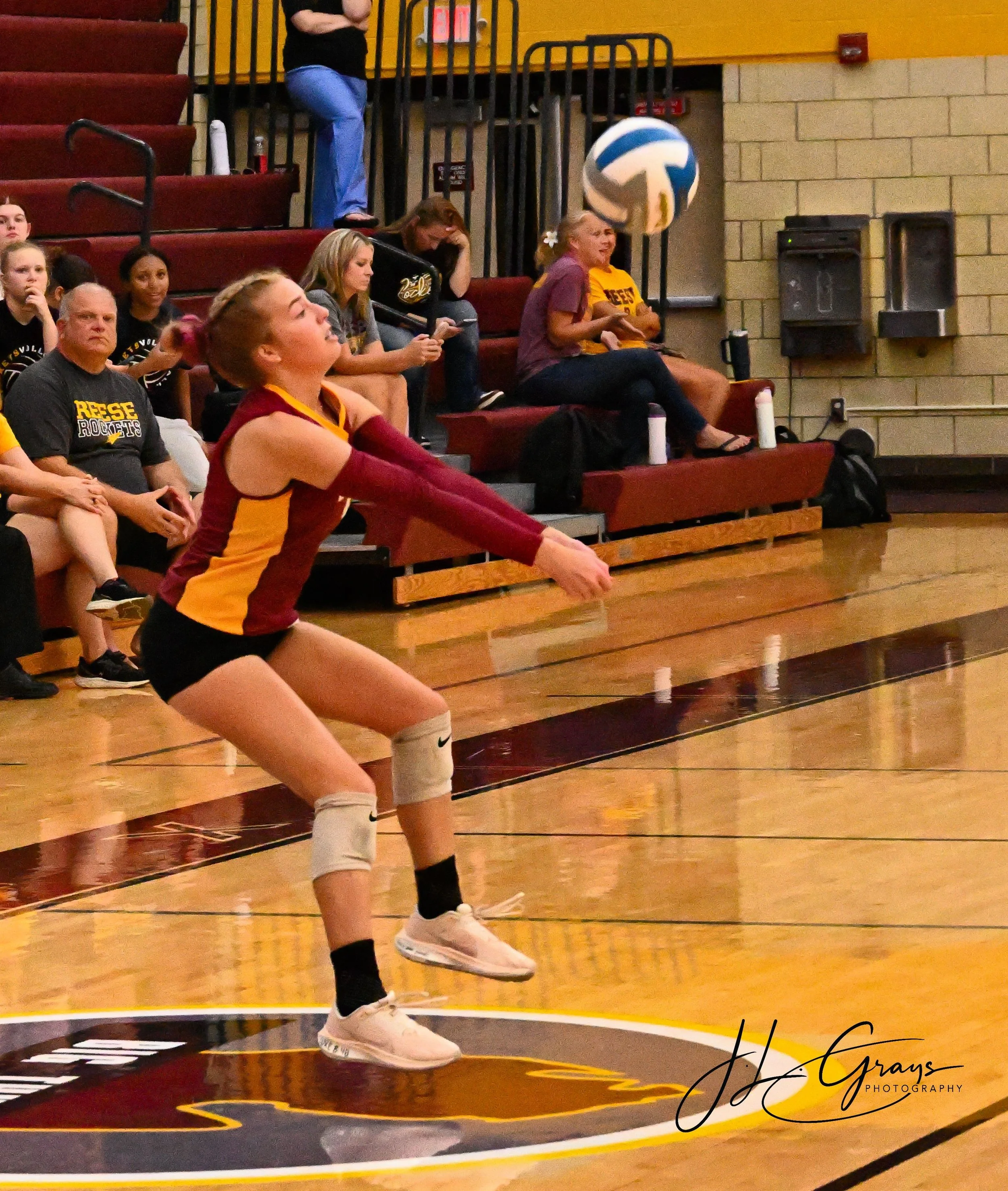 A volleyball player in a maroon and yellow uniform diving to the floor on a gymnasium court, preparing to hit the ball. Spectators sit on the bleachers behind and around the player, with some watching the game and one woman smiling. The gym has a yel