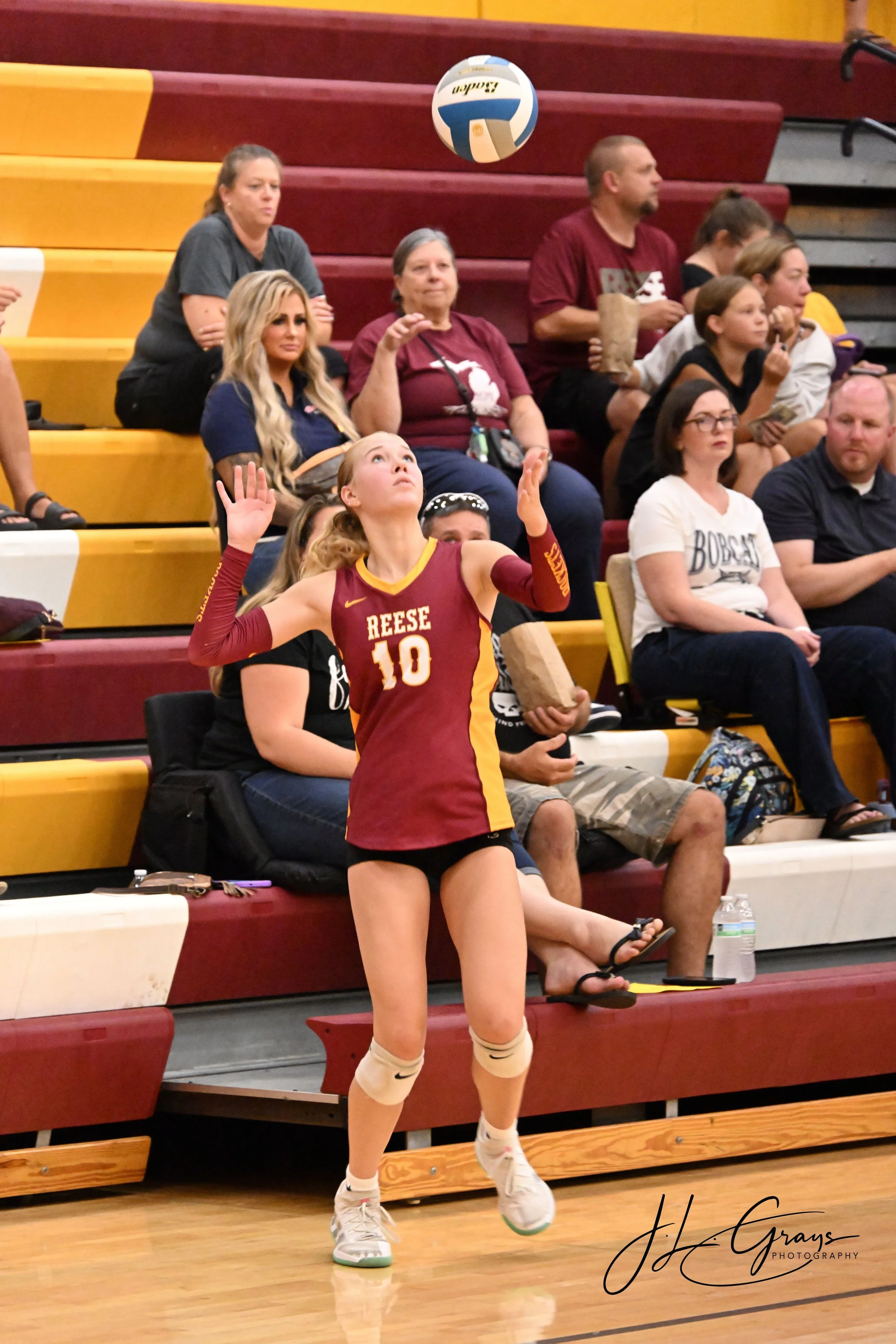 A female volleyball player in a maroon and yellow uniform with the name "Reese" and number 10, is preparing to serve or hit the ball during a match in an indoor gymnasium. Behind her, spectators are sitting on red and yellow bleachers, watching the g