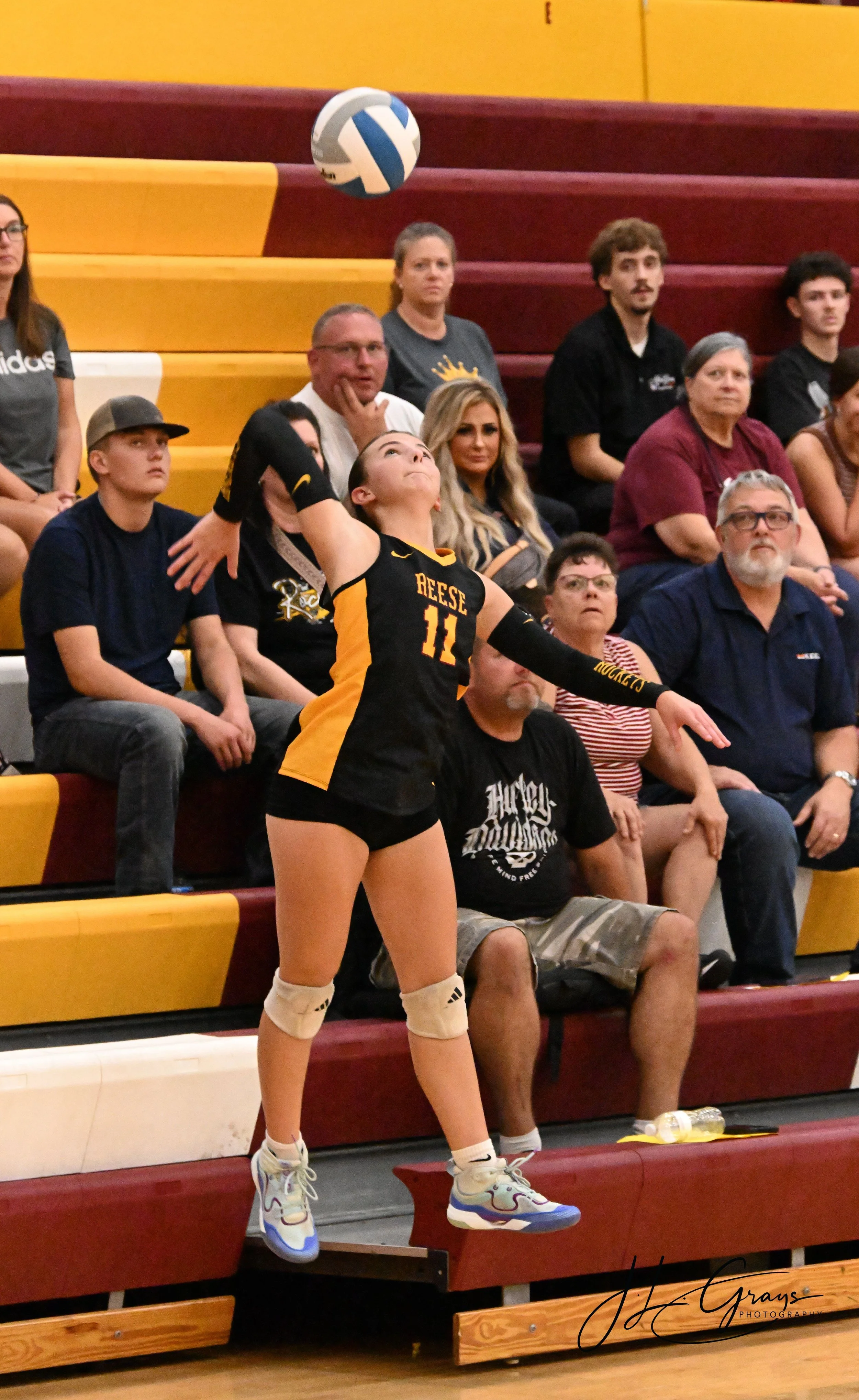 A female volleyball player in a black and yellow uniform jumping to serve the ball during a match in a gymnasium with spectators watching from bleachers.