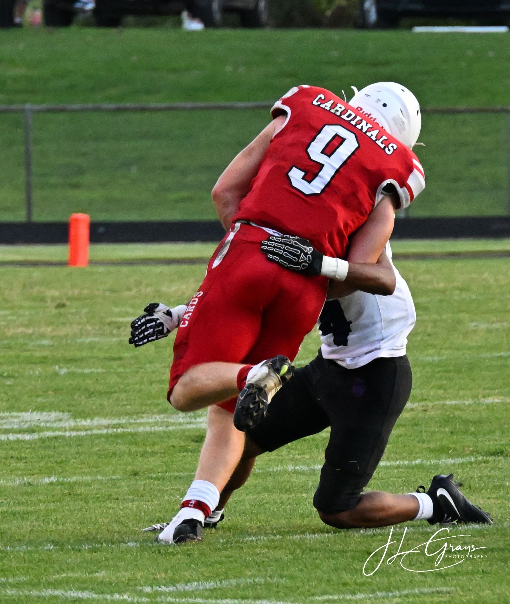 Two football players, one in a red uniform with the number 9, are engaged in a tackle on a football field with green grass. The player in red is grabbing the waist of the other player, who is wearing a white helmet and white uniform, while both are k