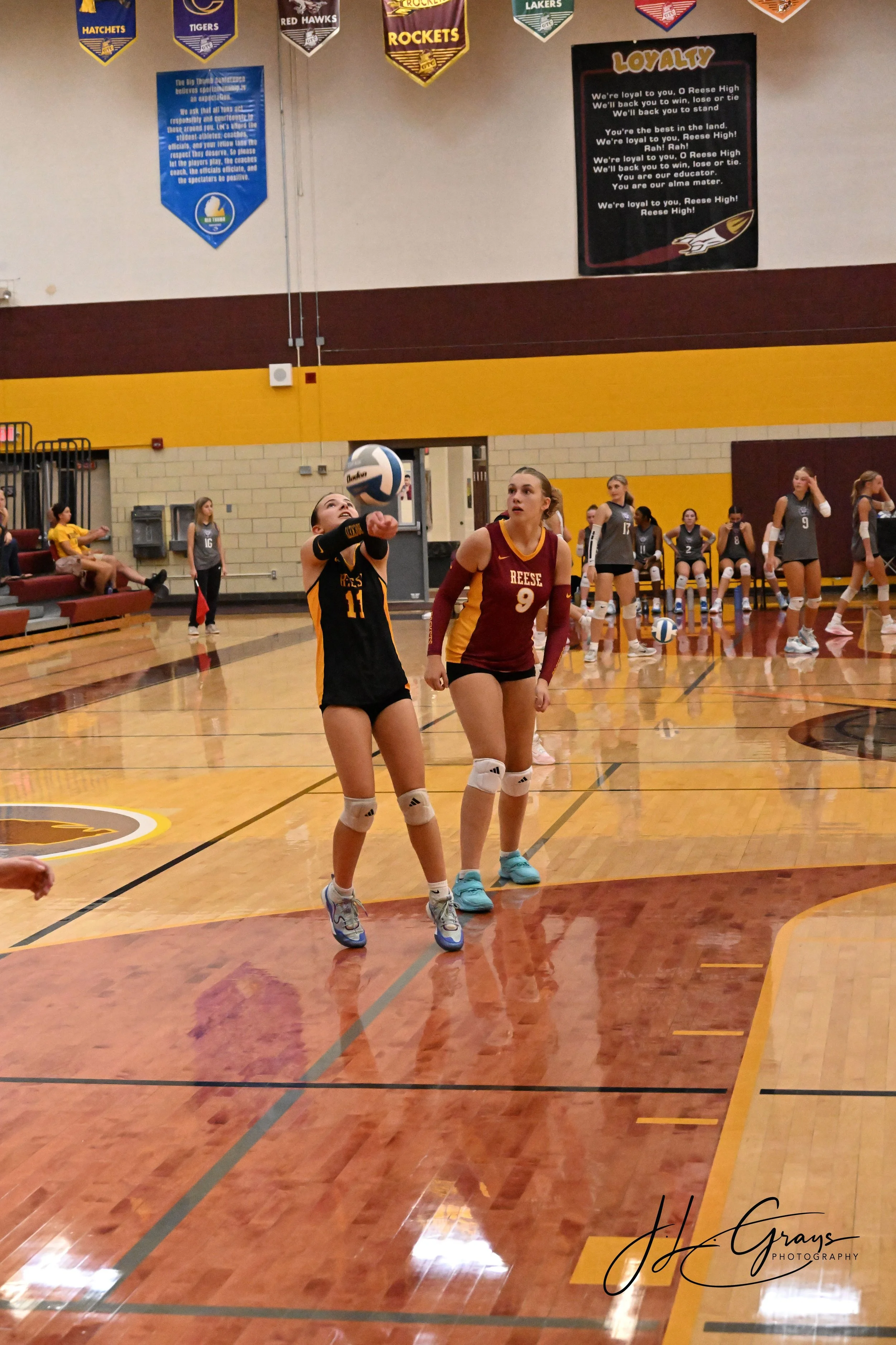 A volleyball game in progress at an indoor gymnasium. Two young women, one in a black and yellow uniform and the other in maroon and yellow, are preparing for a serve. Several players are seated on the bench and others are in the background, some sta