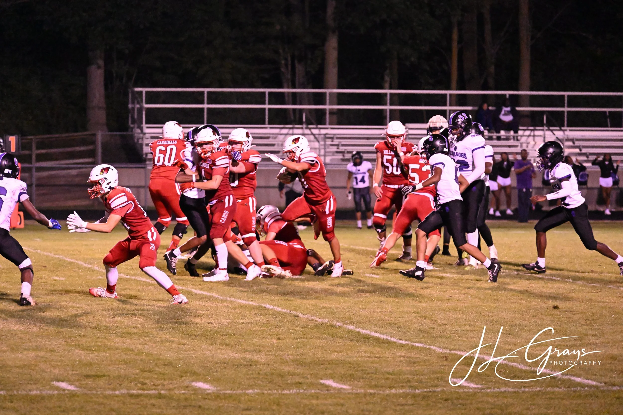 A football game at night with players in red and white uniforms and black and white uniforms on a grass field. Some players are tackling, running, or standing. Spectators are visible in the background sitting on bleachers.