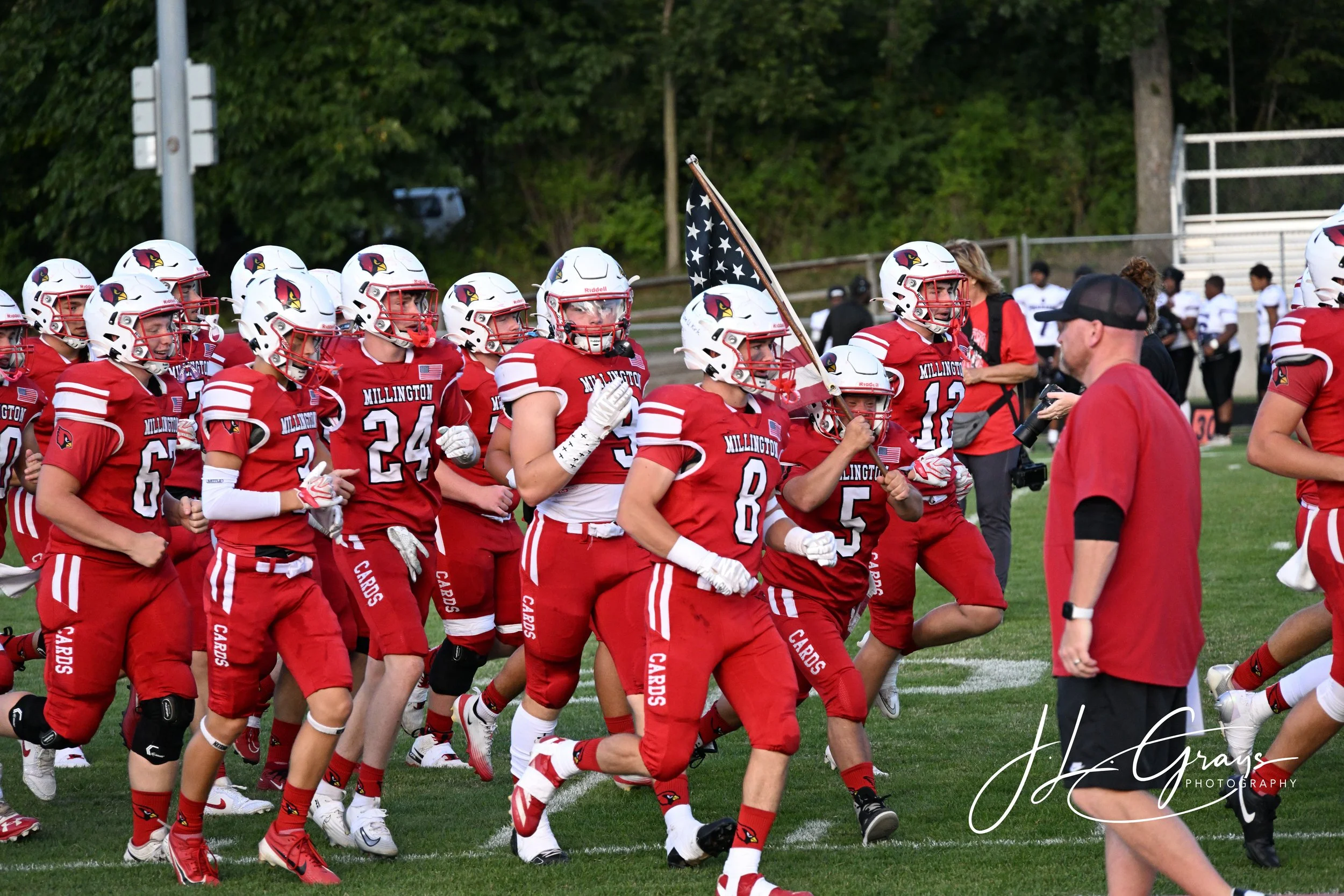 A group of football players in red and white uniforms are jogging onto the field, with some holding a flag. They are wearing helmets and are led by a coach or team staff member in a black cap and glasses. The background shows a grassy field, trees, a