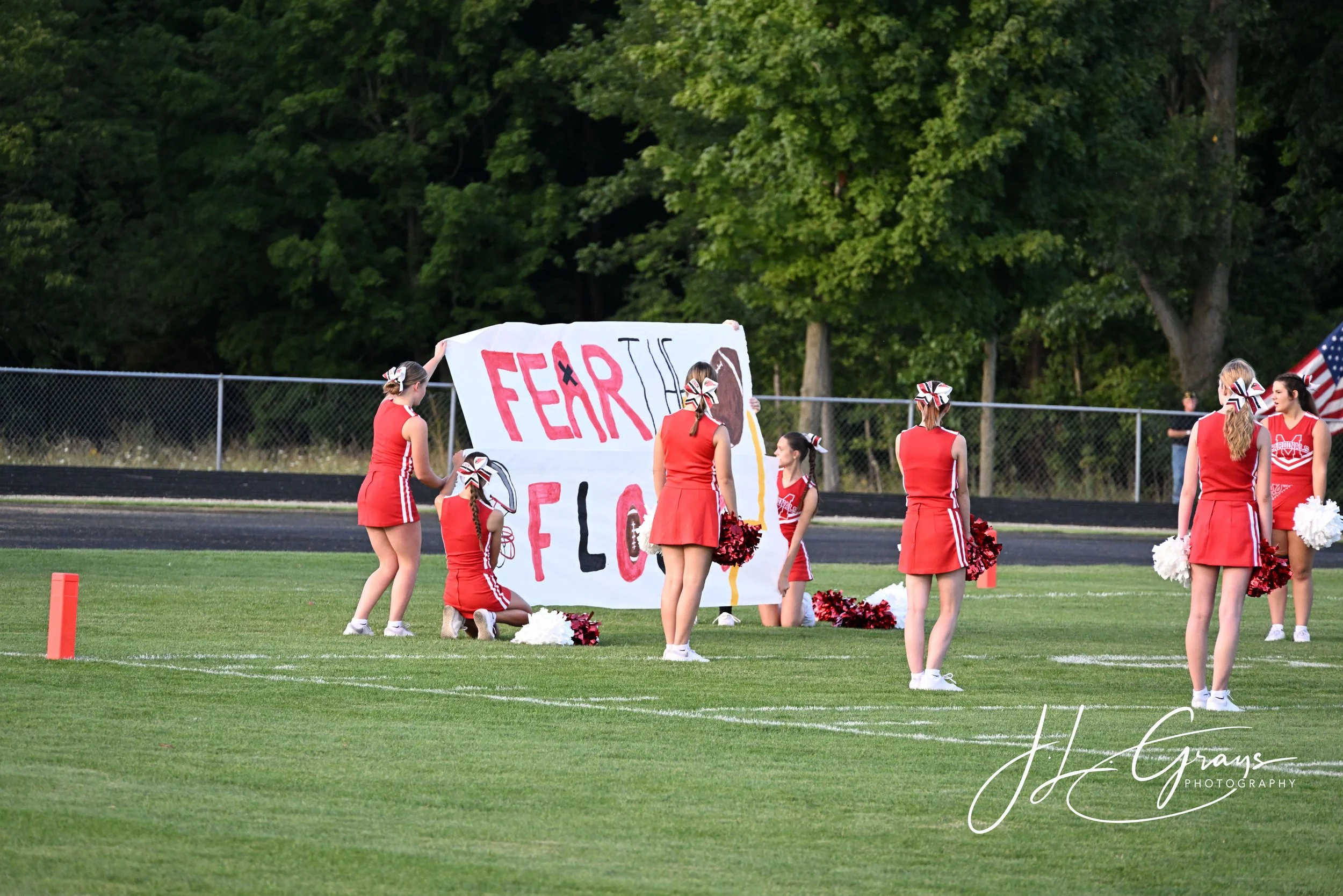 High school cheerleaders in red uniforms and bows, holding pom-poms, standing on a football field during daytime. They are gathered around a large handmade sign that says 'FEAR THE FLOCK' in red and black letters. The background features green trees 