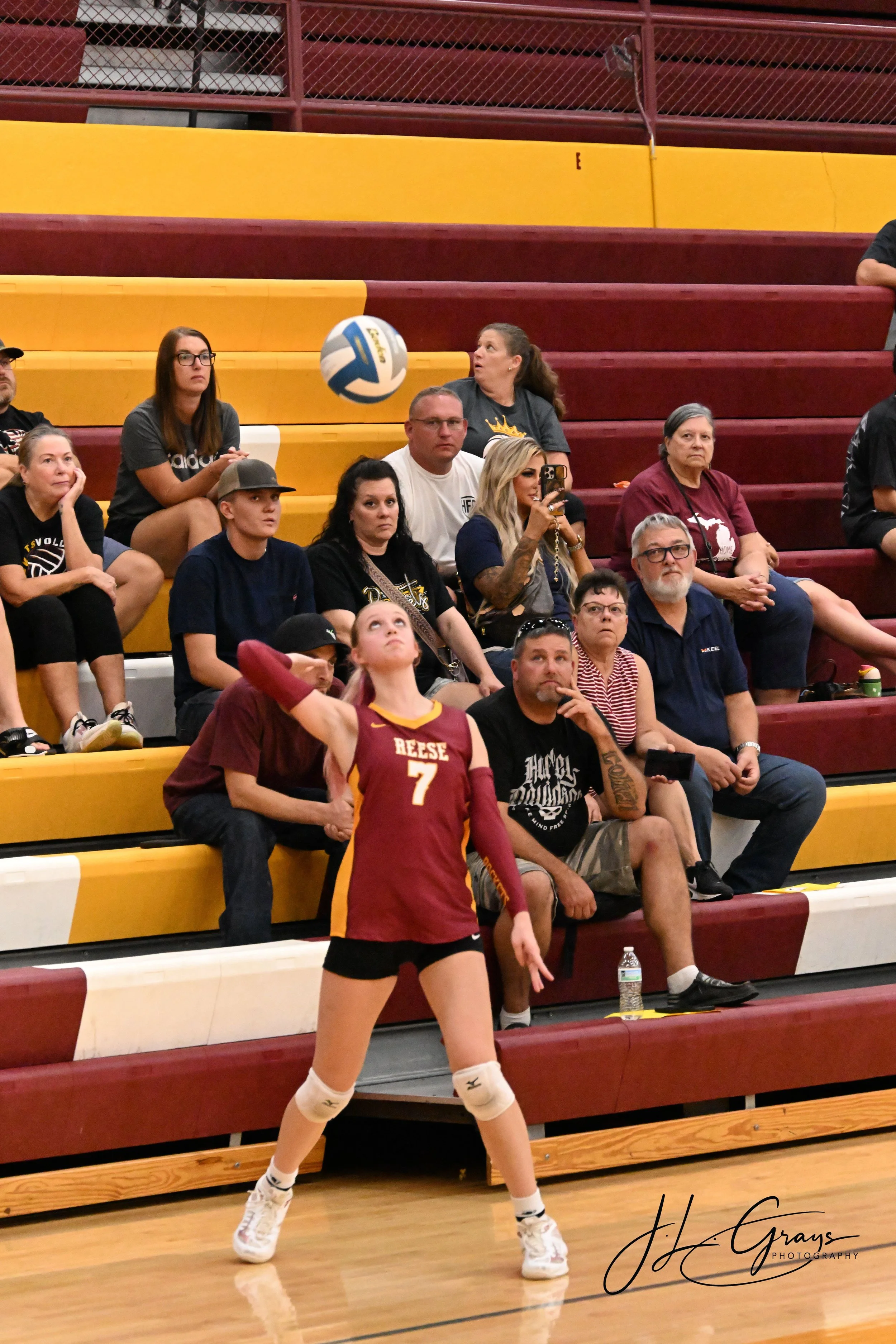 A volleyball player in a maroon and yellow uniform standing on the court with her right arm raised, while spectators watch a volleyball game from the bleachers in the background.