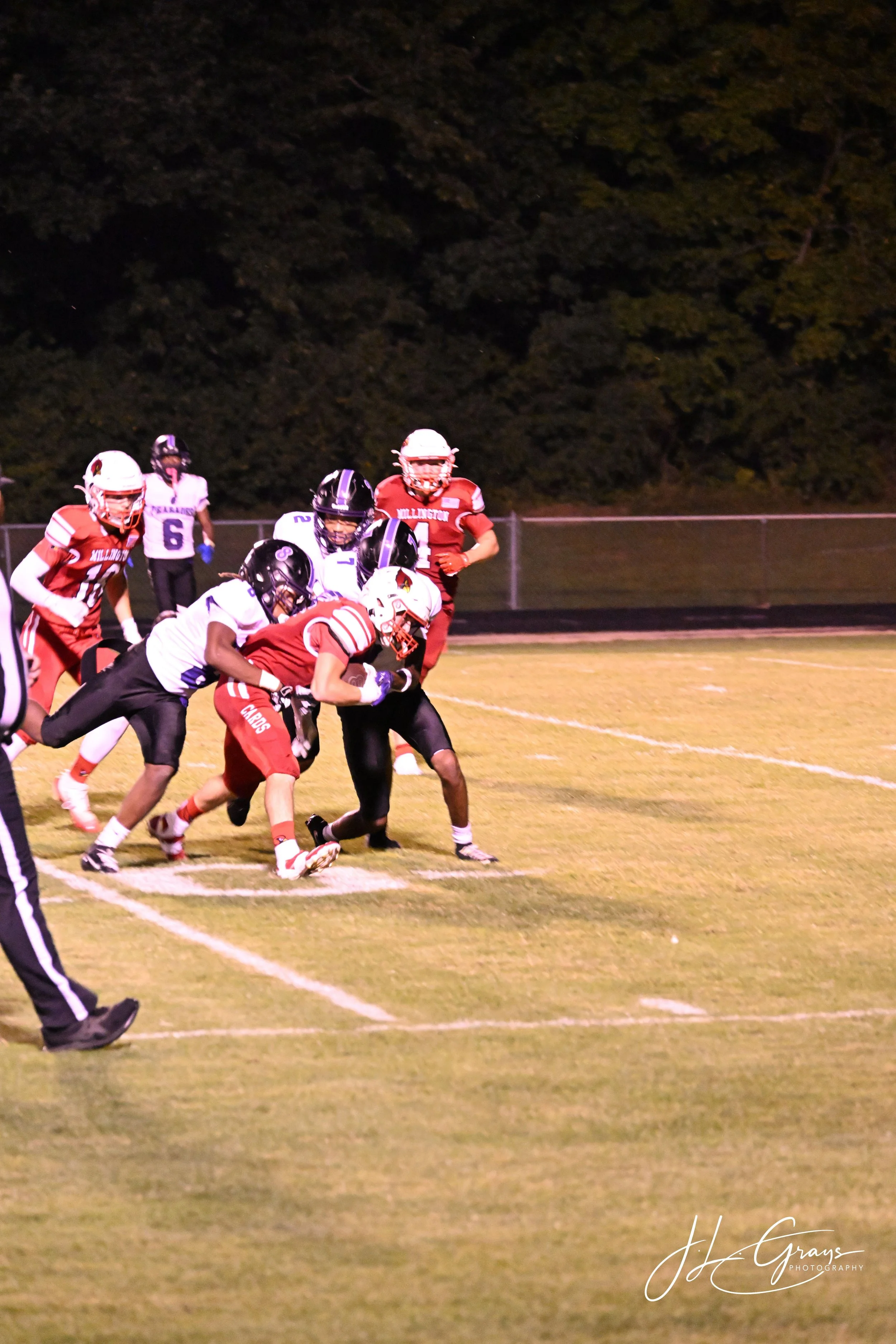 A football game at night showing players in red and white uniforms competing on the field, with the game in progress, and dark trees in the background.