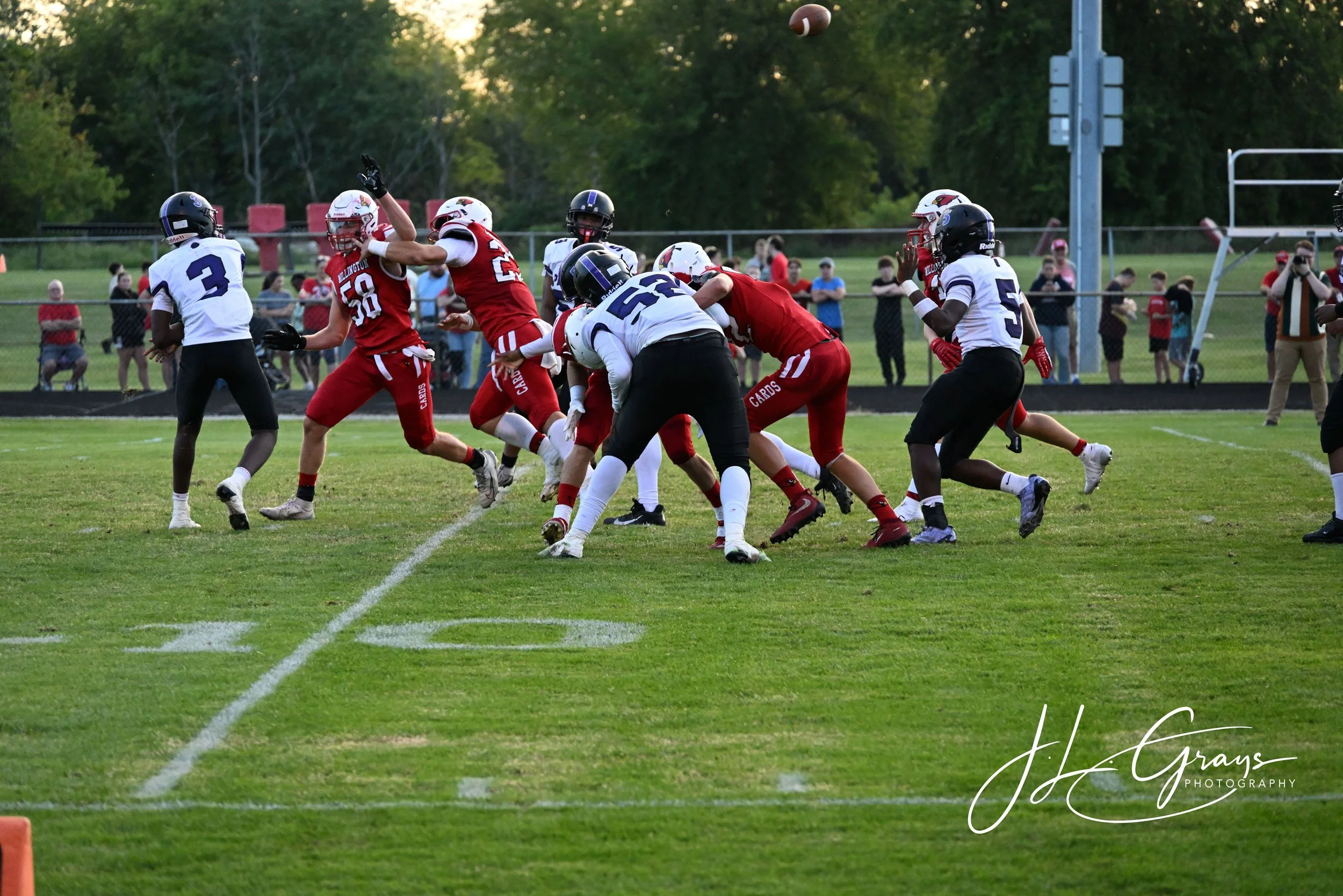 A youth football game showing players in action on the field, with some players tackling and others running with the ball. Spectators watch from the sidelines.
