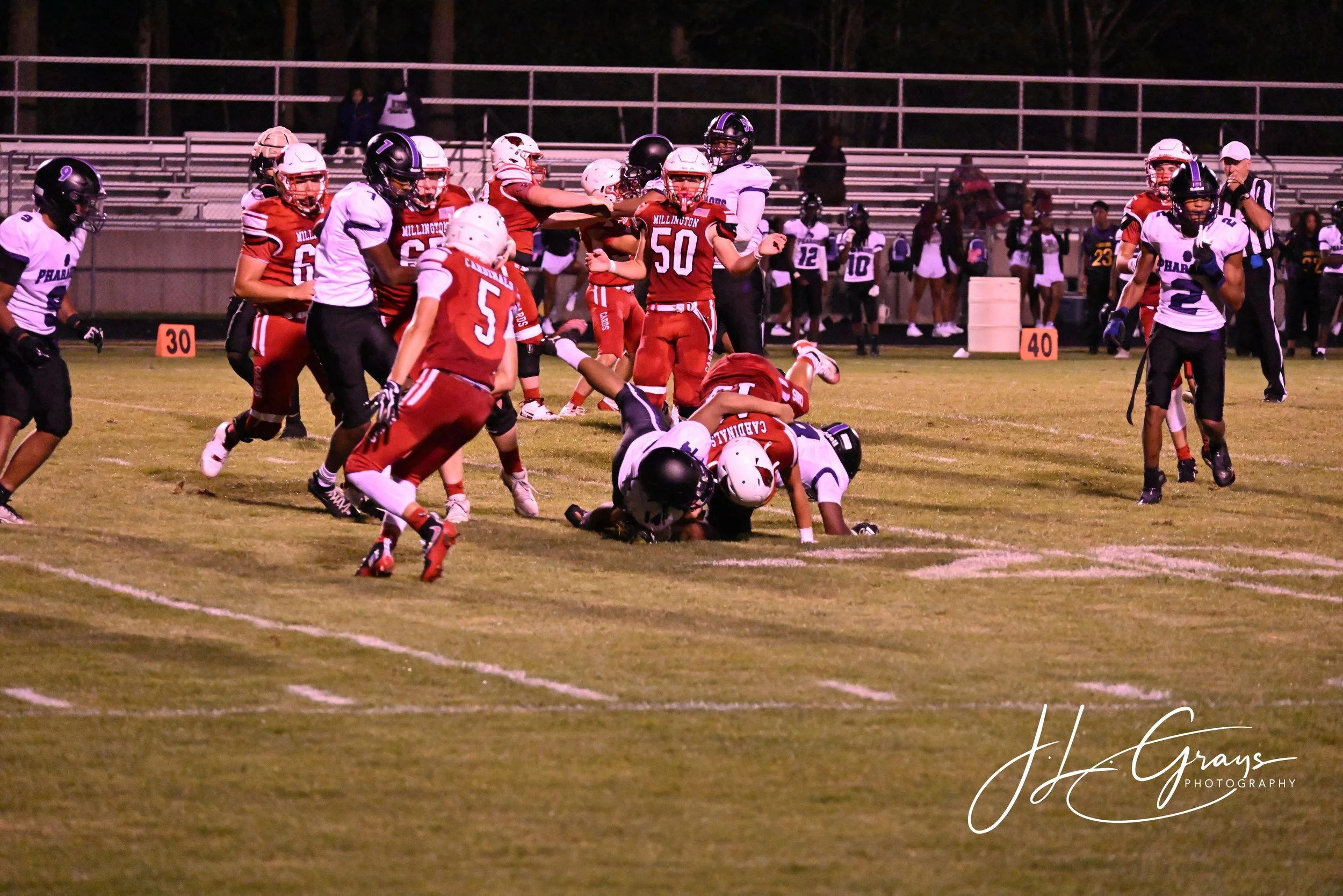 A football game in progress with players in red and white uniforms on the field, some tackling and others running, while spectators watch from the bleachers at night.