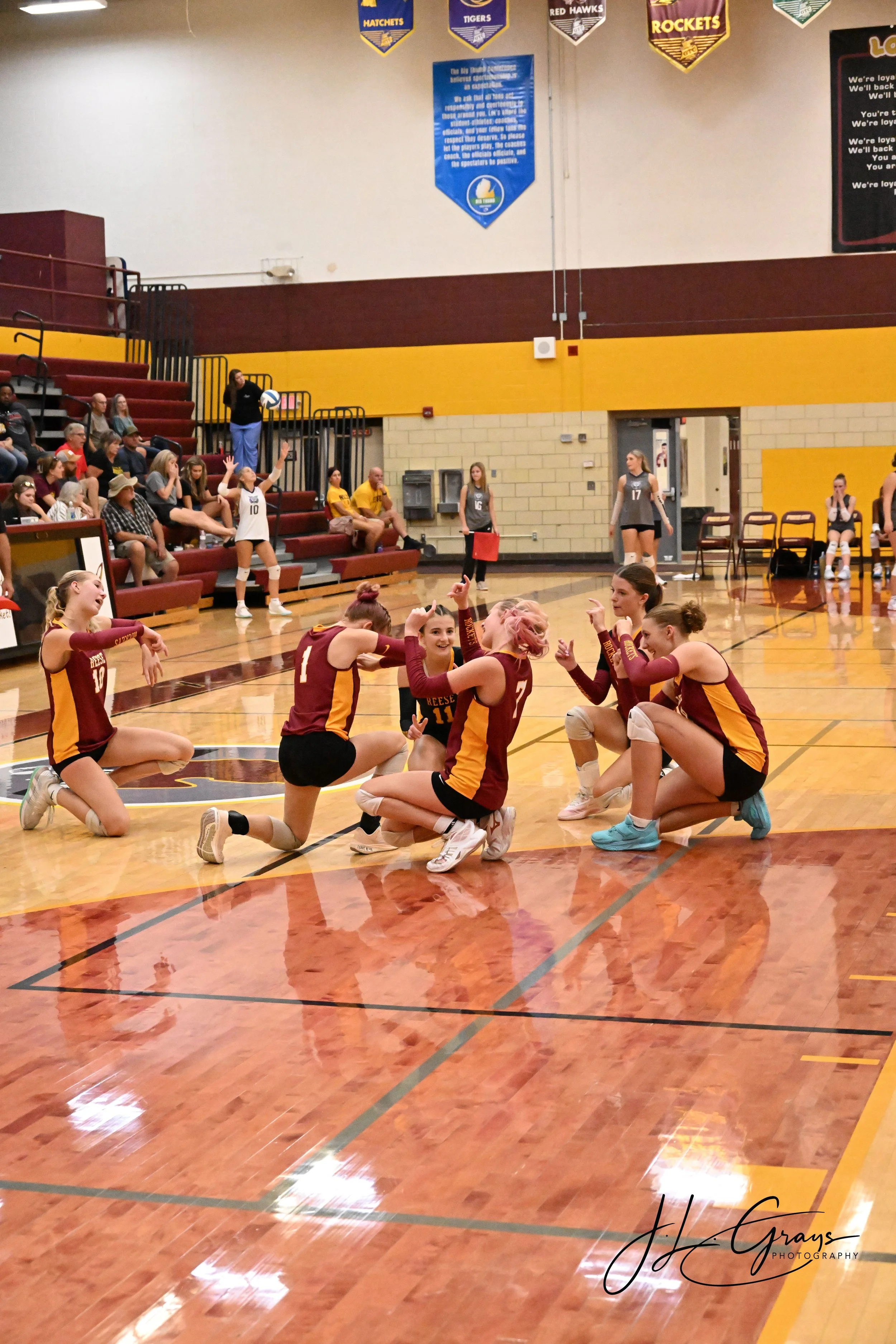 A volleyball team wearing maroon and yellow uniforms is celebrating on an indoor court, with some players kneeling and others standing. Spectators are seated in the bleachers, some clapping and others watching. Banners hang from the gym ceiling, and 