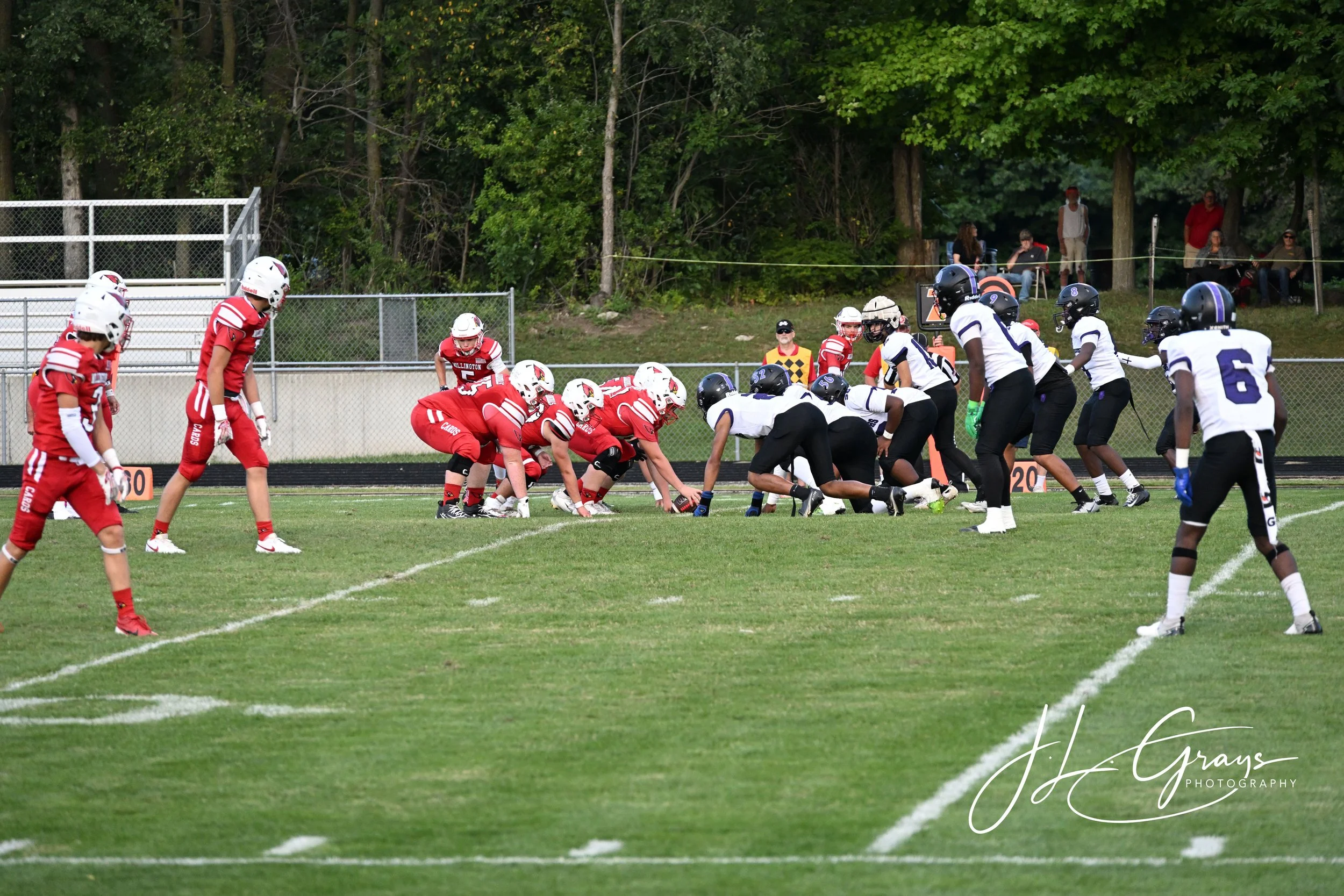 American football game with players in red and white uniforms and black helmets on the field, with spectators in the background.