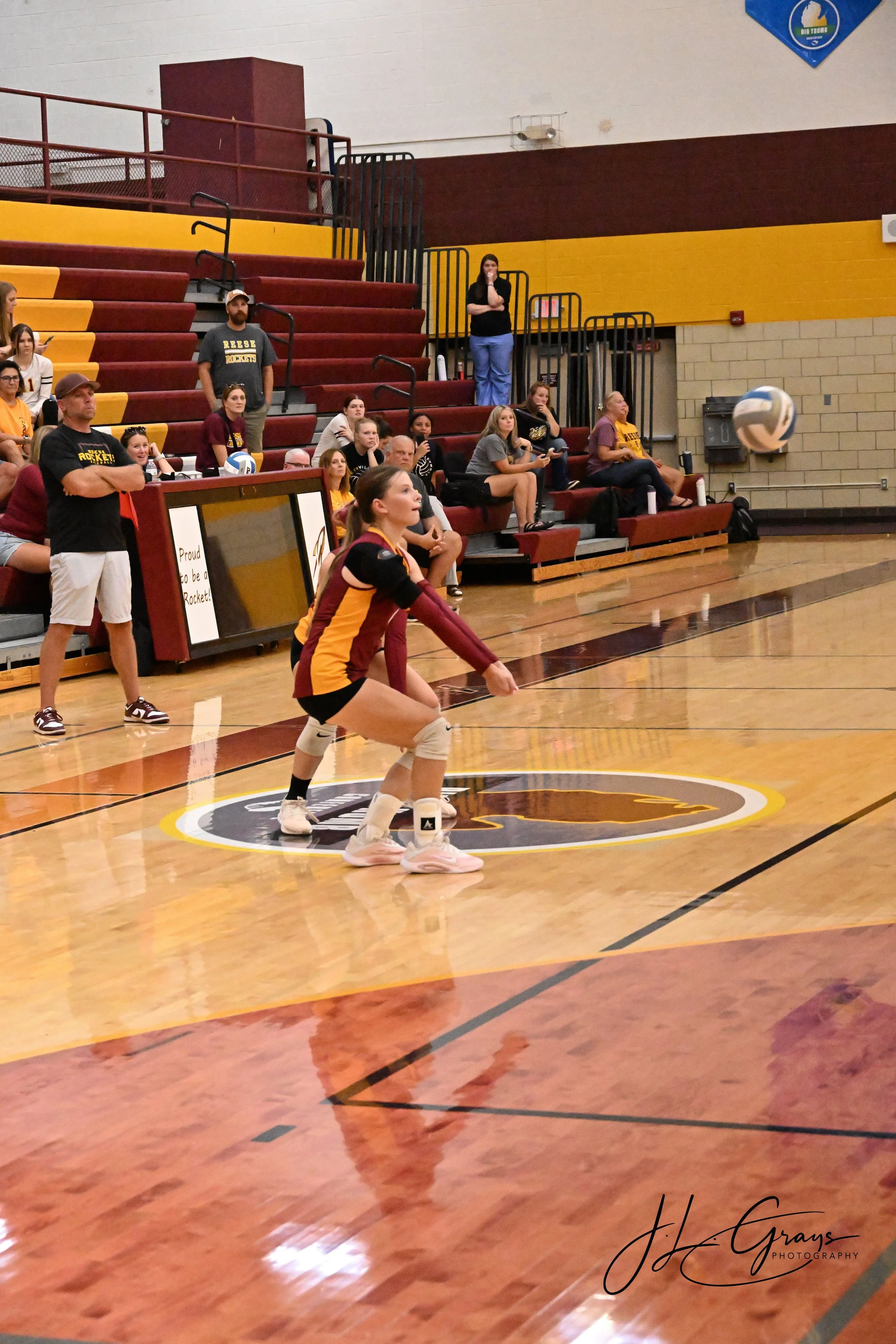 A volleyball match in progress at an indoor gymnasium. A female player in a maroon and yellow uniform is crouched, ready to receive the ball. Spectators and coaches are sitting on bleachers watching the game, with some watching intently and others en