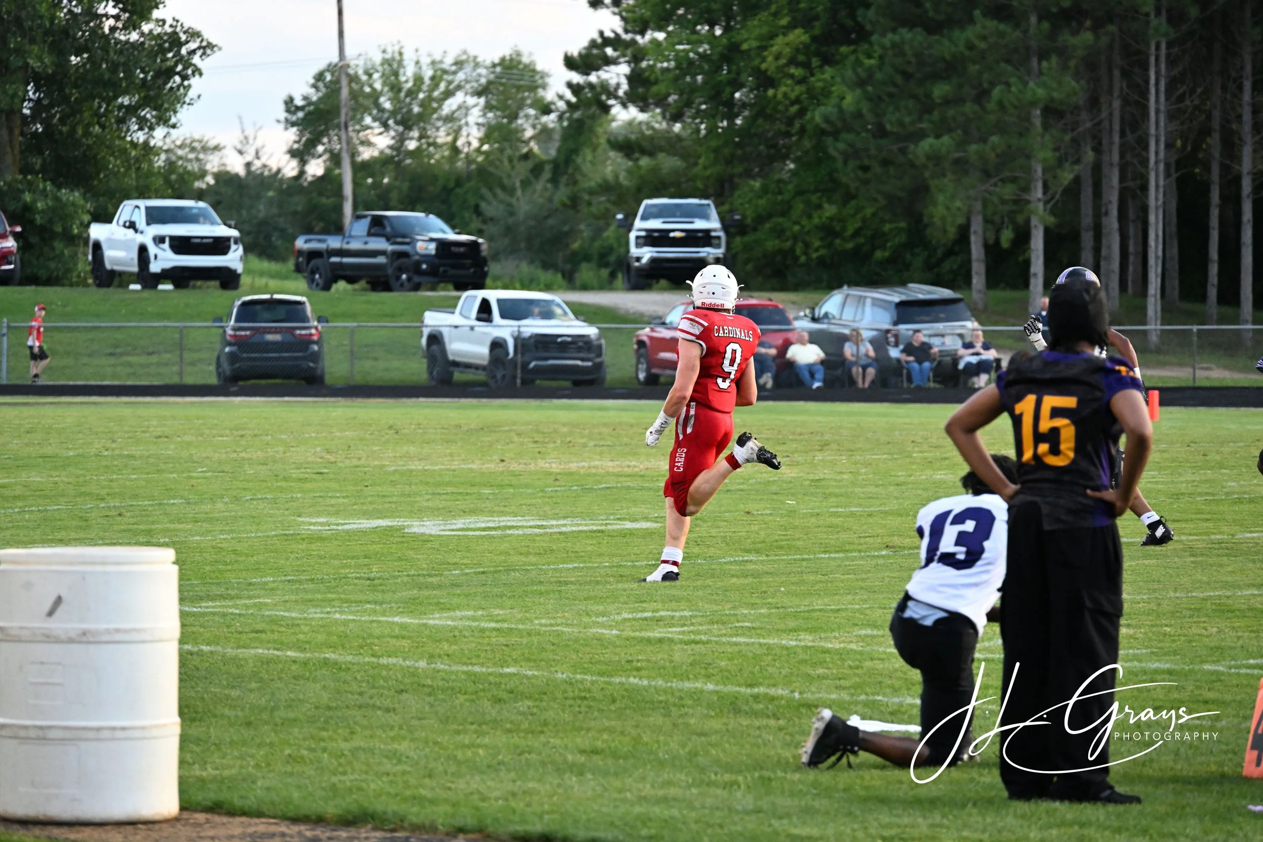 A football game with players in red and black uniforms on the field. In the background, there are spectators sitting on benches and parked cars behind a fence, with trees and a cloudy sky overhead.