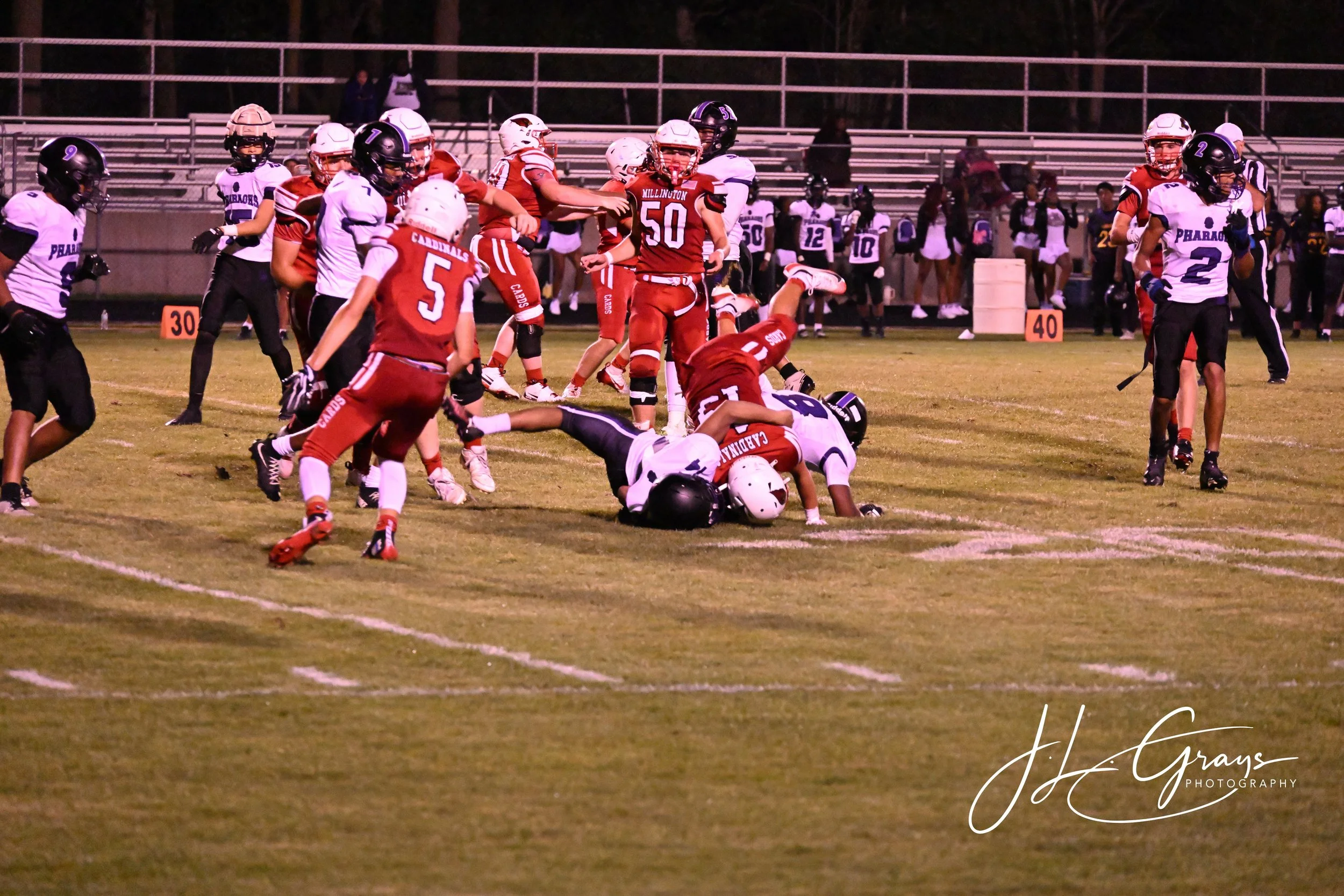 A youth football game at night with players in red and white uniforms on the field. One player in red is on the ground tackled by a white-uniformed player. Other players are standing or moving around, with some on the sidelines and spectators in the 