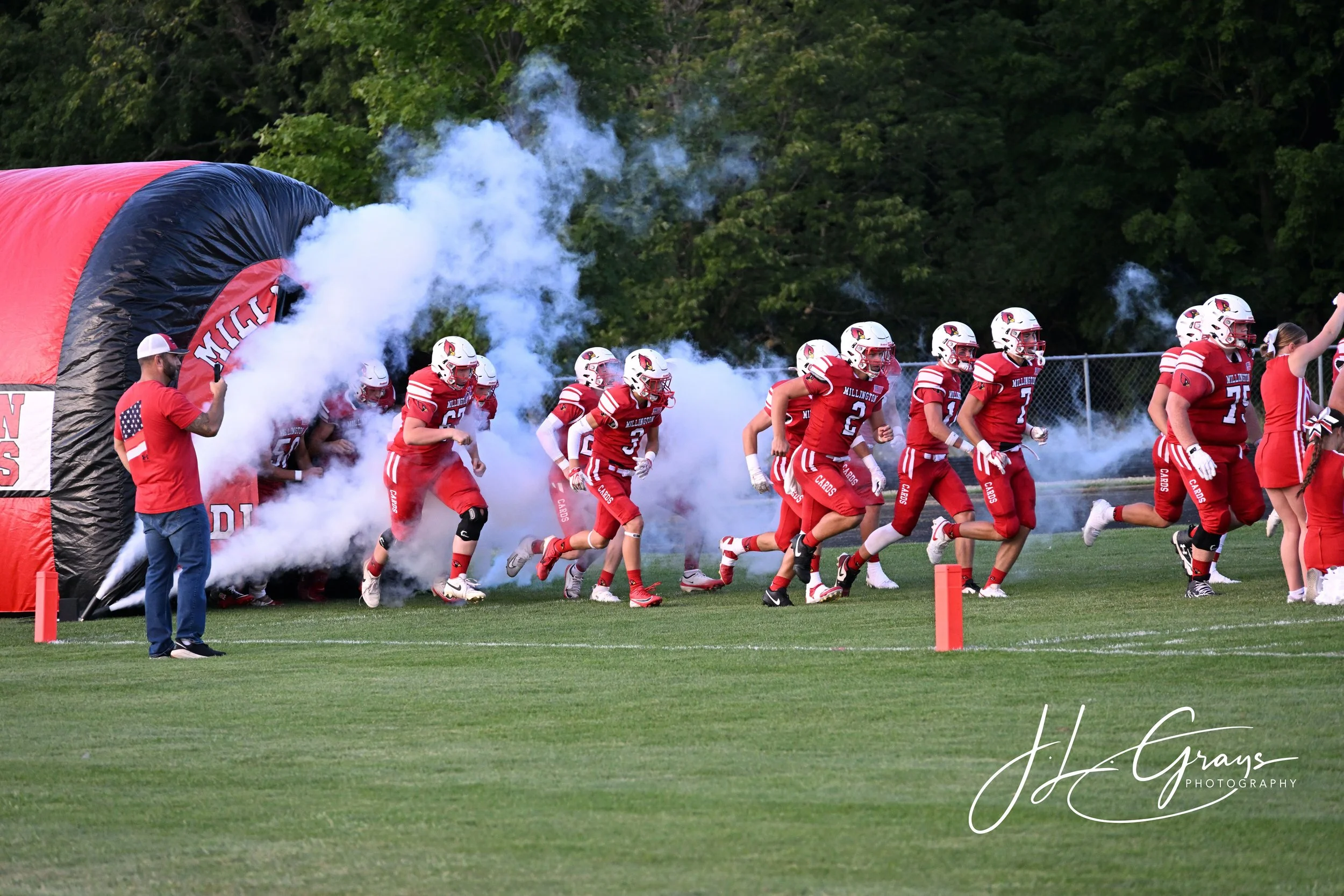 A group of American football players in red uniforms running onto the field from a tunnel with smoke, led by a coach or team staff member holding a whistle, during a daytime game. The players are wearing helmets and football gear, and a chain-link fe