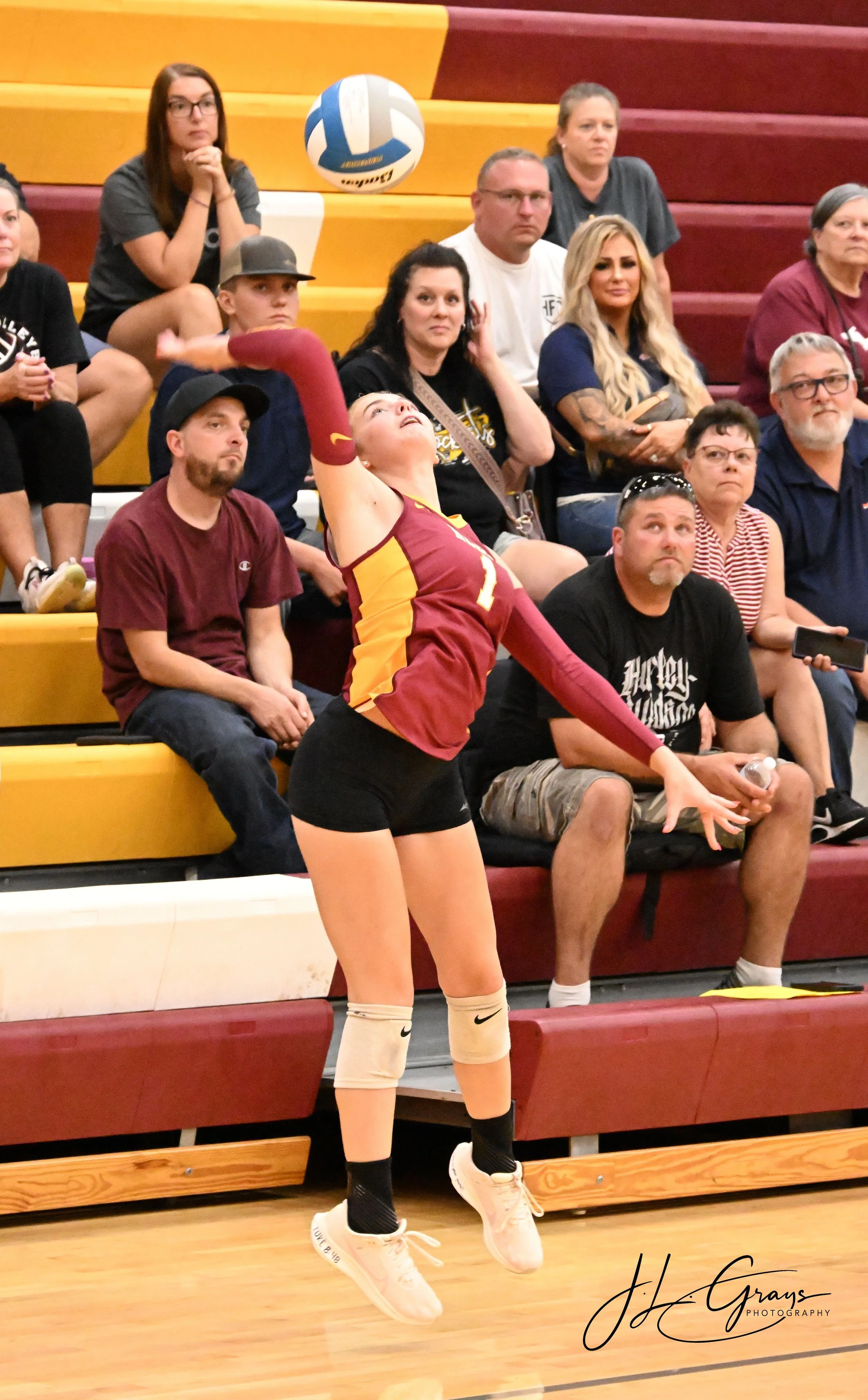 A female volleyball player in a maroon and yellow uniform serves during a match, with spectators watching from the bleachers behind her.