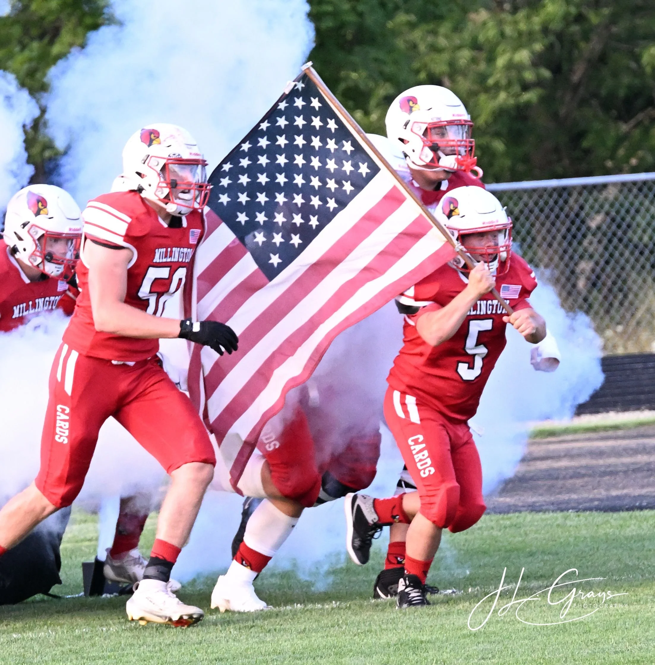 Members of a football team, dressed in red uniforms with a cardinal logo on their helmets, running onto the field carrying an American flag with a blue field of white stars and red and white stripes, as smoke billows behind them.