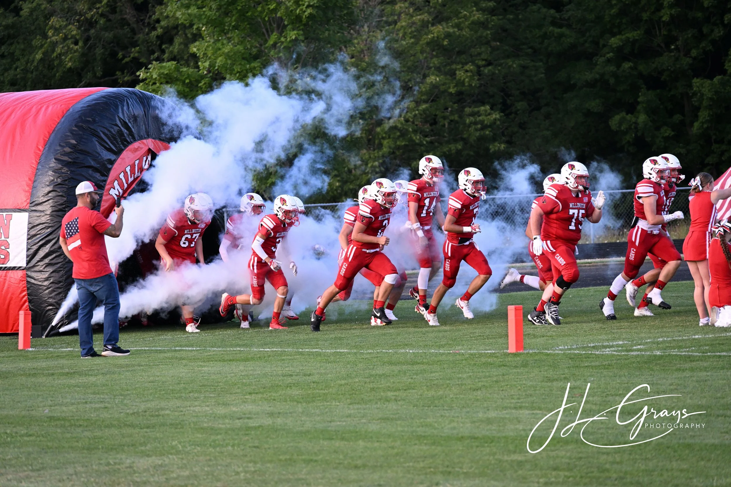 A group of football players in red uniforms run onto the field through smoke, emerging from a tunnel with a red and black cover, as an adult man holding a smoke device watches.