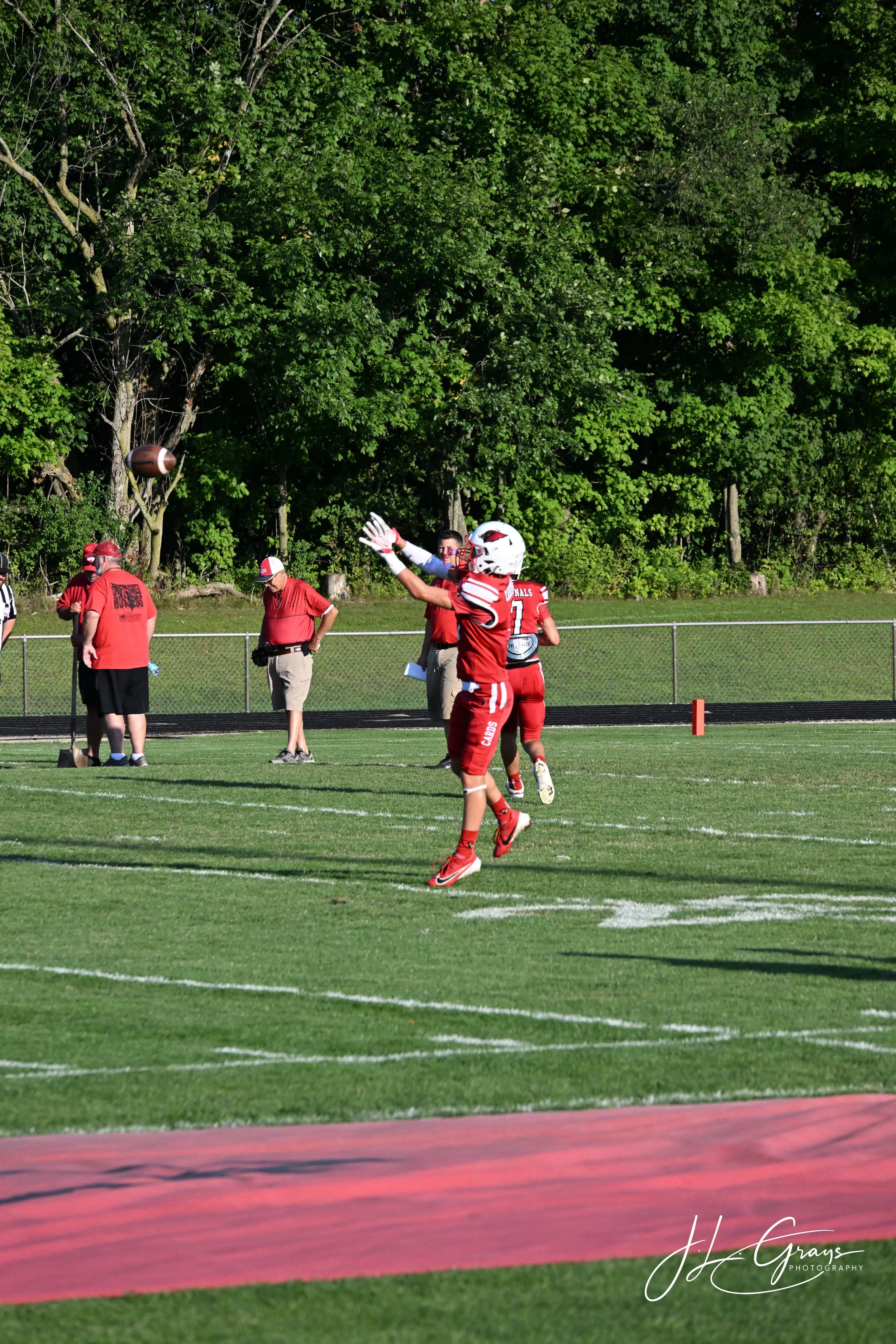 American football players in red uniforms warm up on the field before a game, with trees in the background.